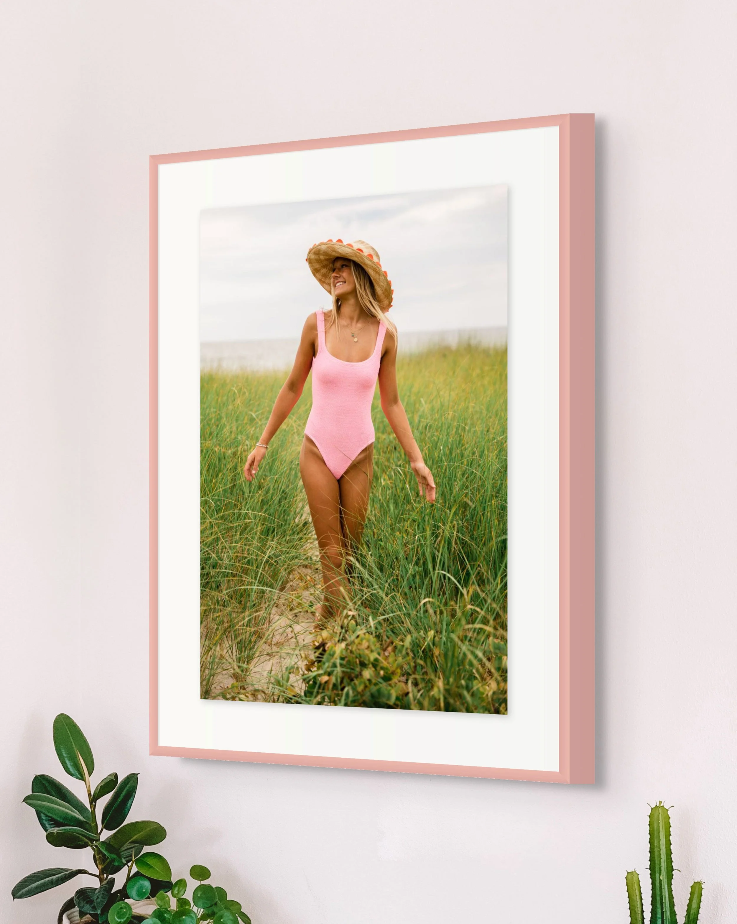 Framed photo of a woman in a pink swimsuit walking through a grassy field, wearing a large straw hat with orange accents, near a beach.