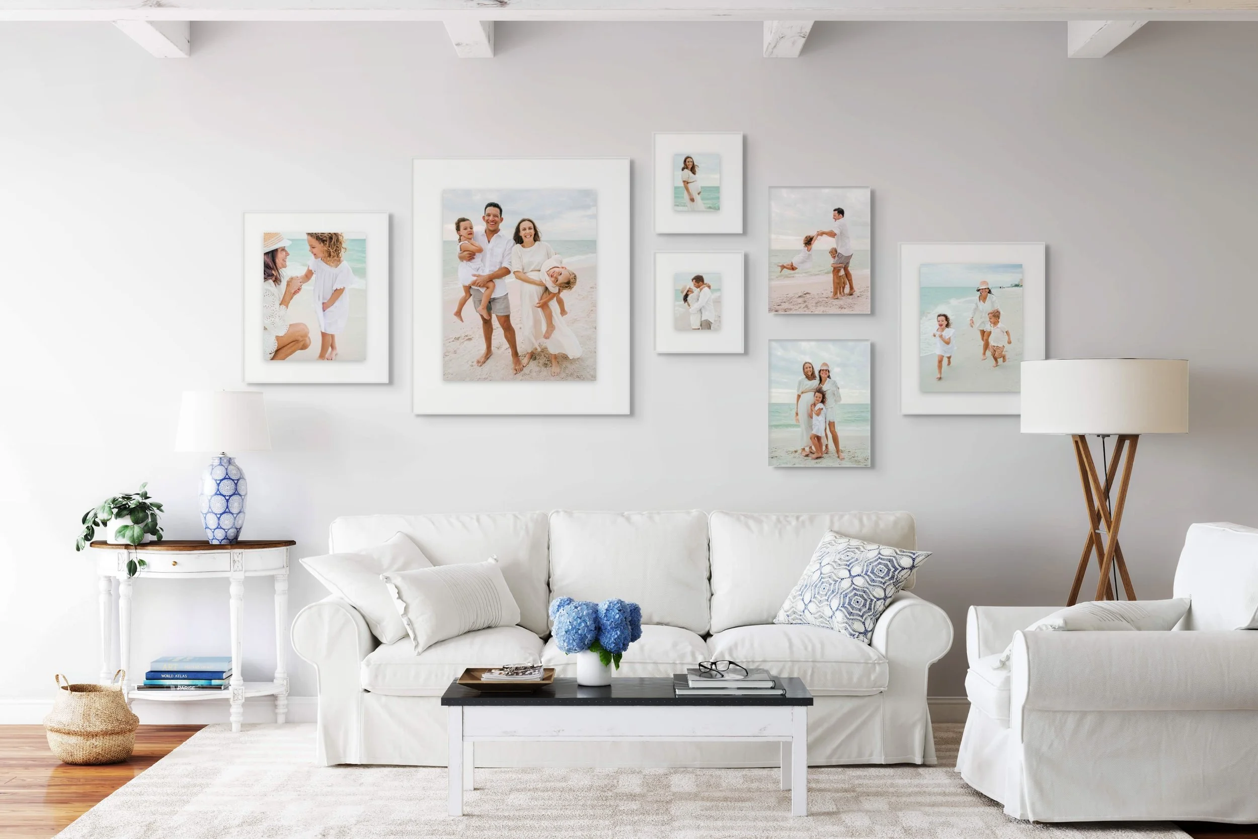A bright living room with white sofa, armchair, and a coffee table with blue flowers. The wall above features a collage of framed family beach photos.