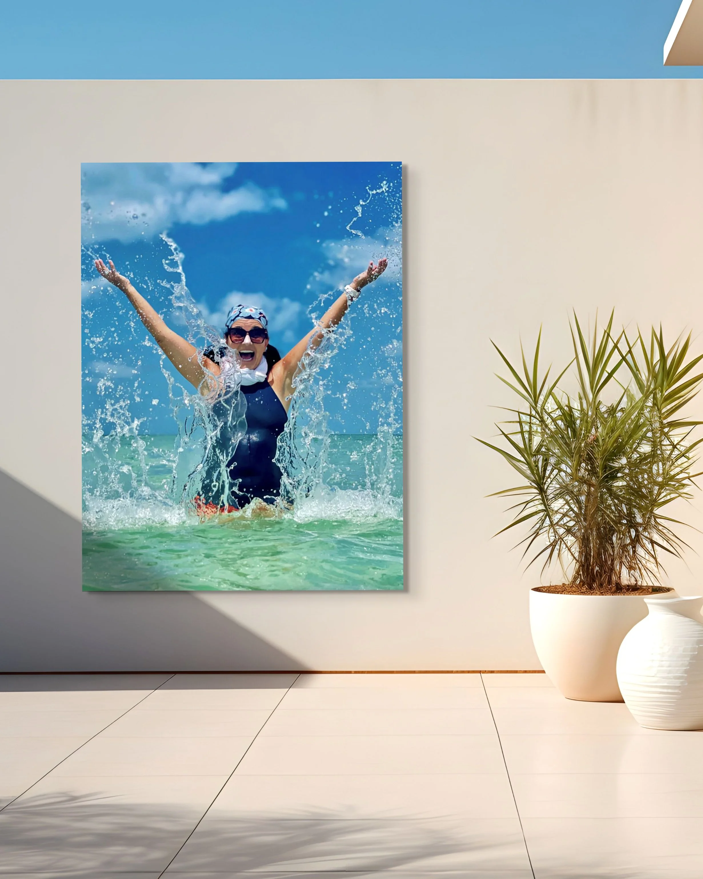 A framed photograph of a woman swimming in the ocean, smiling with arms raised, wearing sunglasses and a swim cap, against a cloudy blue sky background, hanging on a light-colored wall next to a potted plant with spiky green leaves.