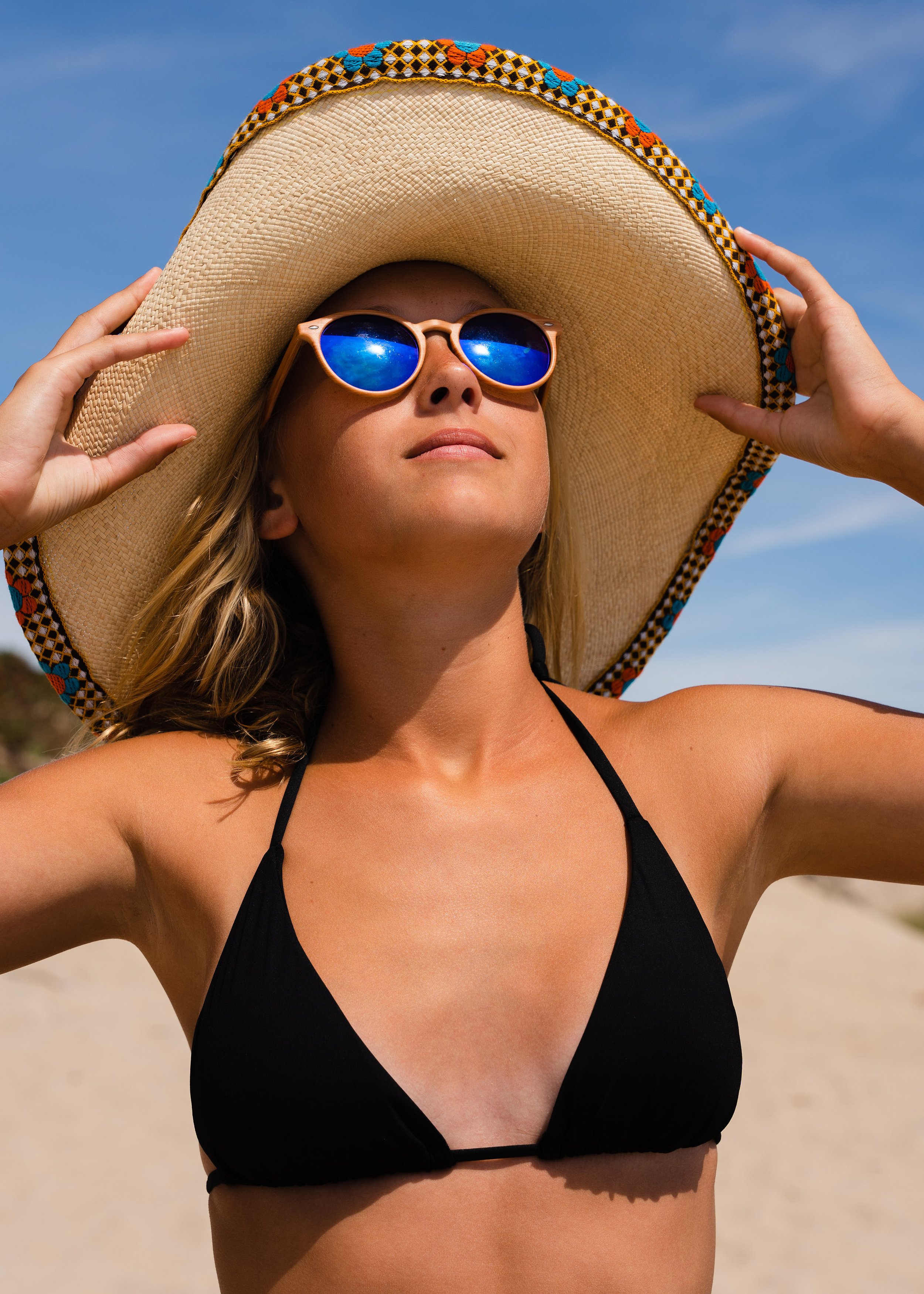 A woman wearing a large sun hat and reflective sunglasses, standing outdoors with a blue sky in the background, dressed in a black bikini top.