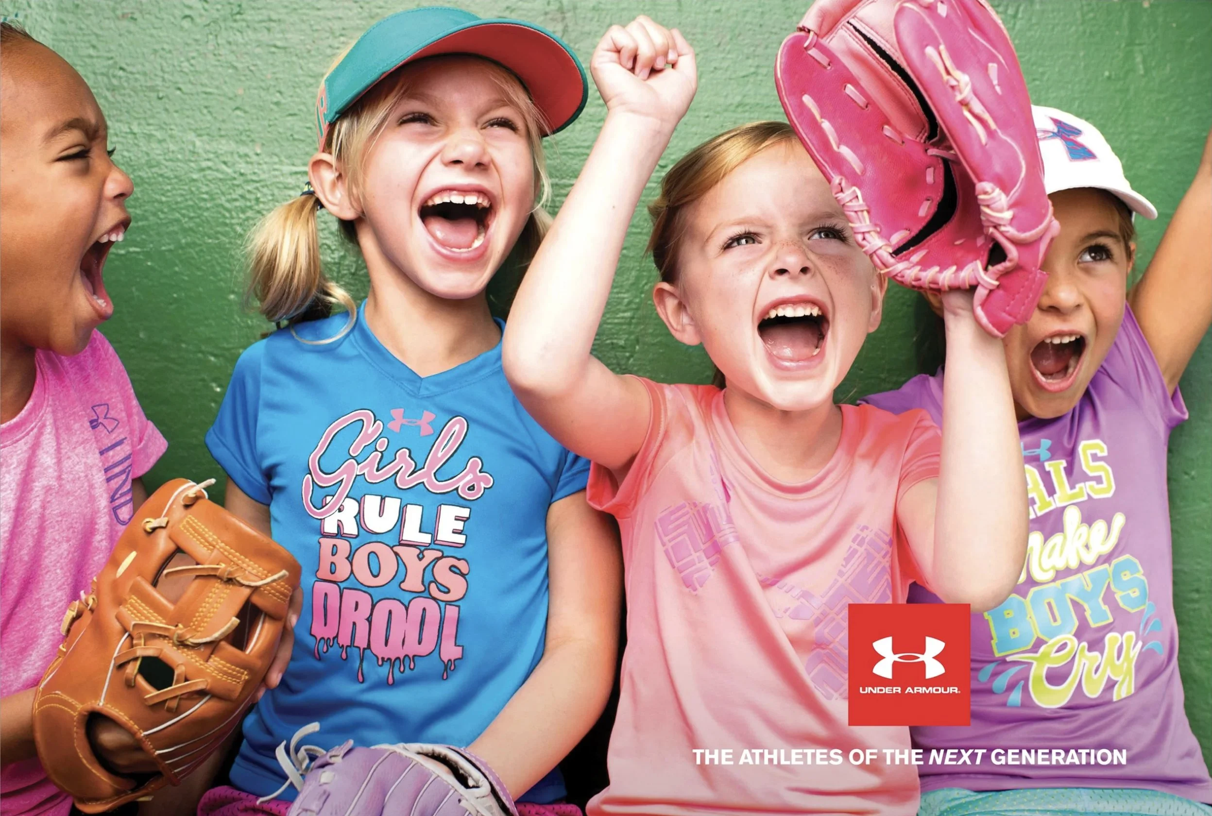 A group of young girls at a sports team photo shoot, wearing colorful athletic shirts, gardening gloves, baseball caps, and baseball mitts, smiling and laughing against a green background.