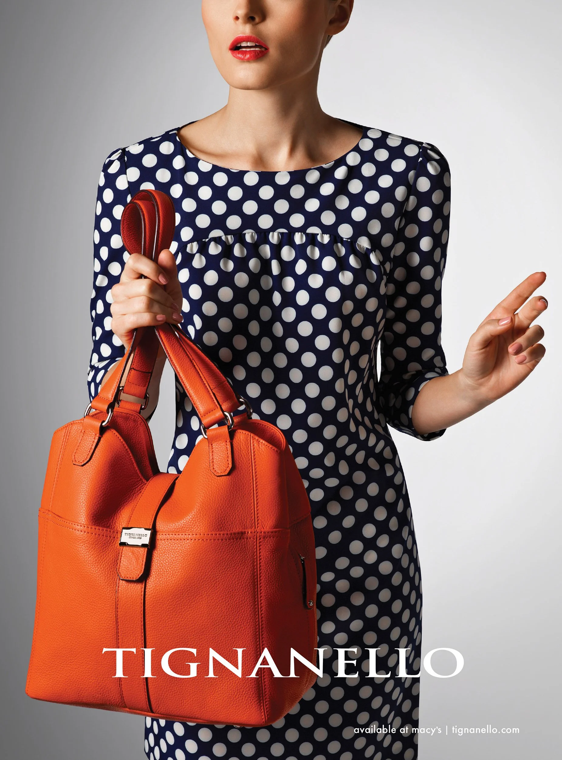 A woman is holding an orange handbag with a textured surface and silver accents, wearing a navy blue dress with white polka dots, and red lipstick. The image promotes Tignanello handbags available at Macy's.