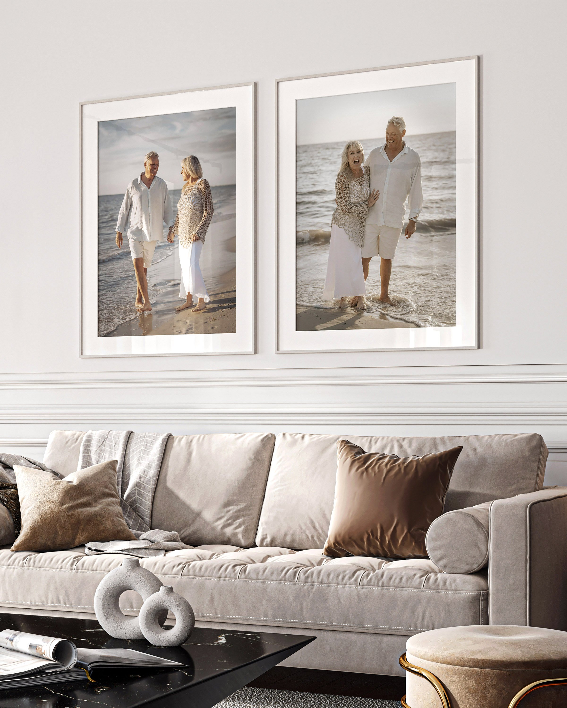 Living room with beige sofa, decorative pillows, black coffee table with white sculptural objects, two modern paintings of couples on the beach, beige ottoman with gold accents.