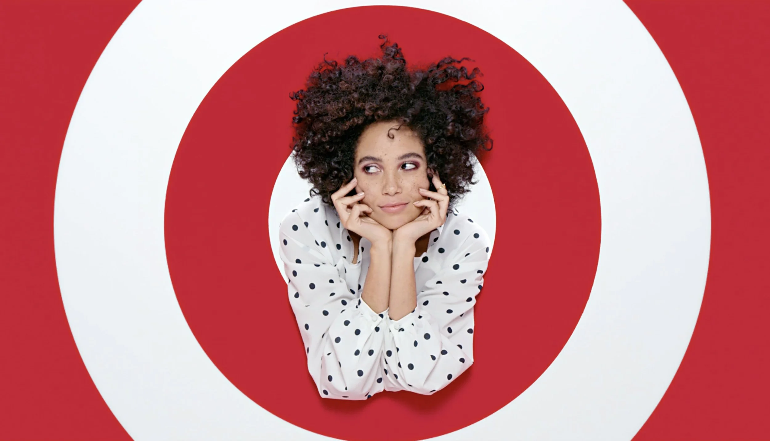 A woman with curly hair and a white polka dot shirt resting her chin on her hands inside the bullseye of a red and white Target logo.