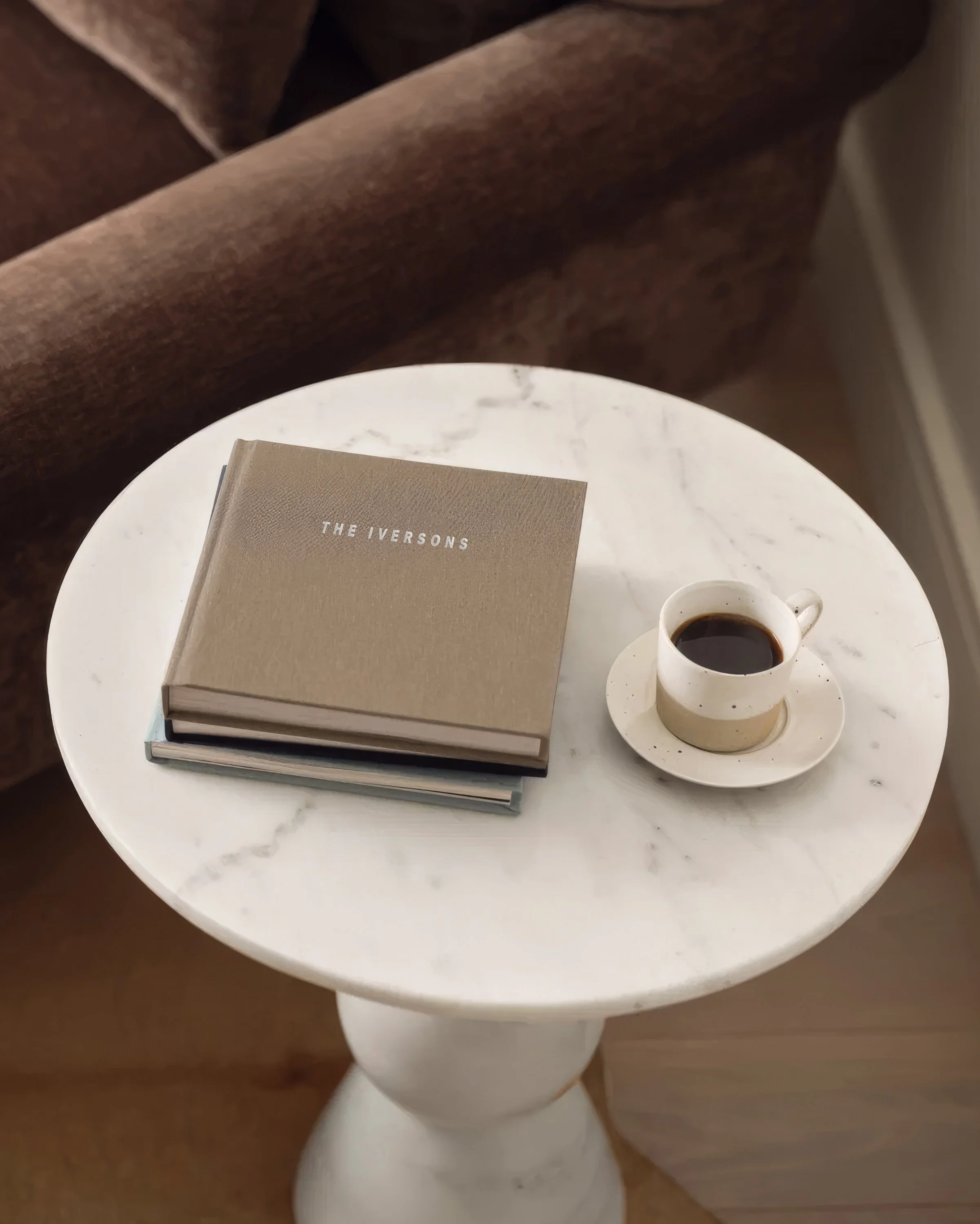 A white round table with a closed book titled 'The Iversons' and a cup of black coffee on a saucer, against a background of a brownUpholstered sofa.