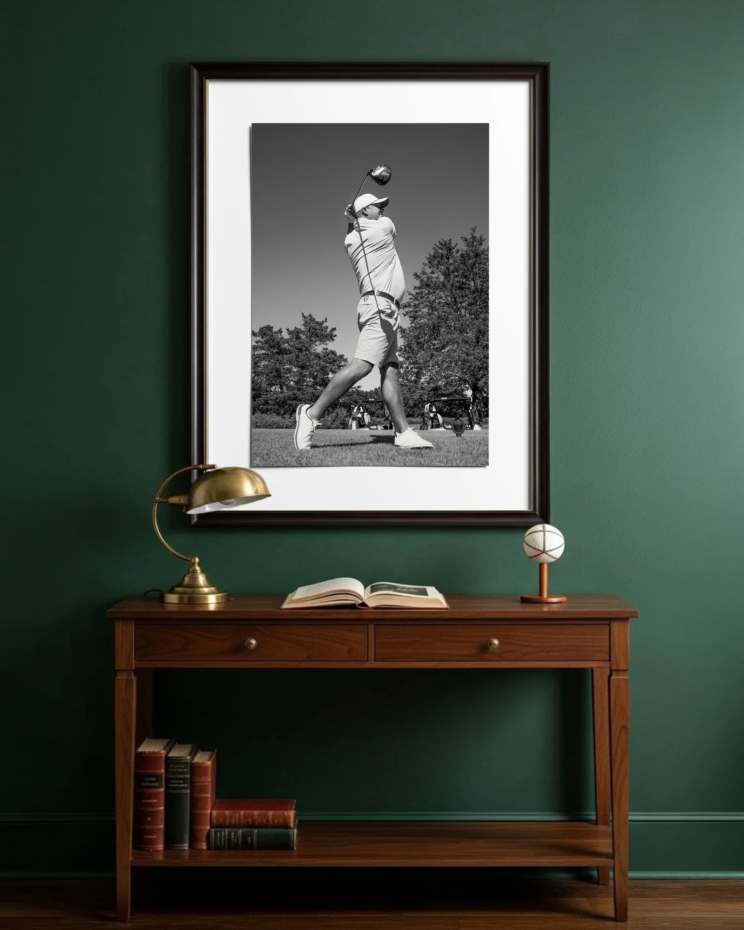 A wooden console table under a framed black-and-white photograph of a male golfer swinging a club. The table holds a gold desk lamp, an open book, and decorative spherical objects. To the left of the table on the floor are stacked books.