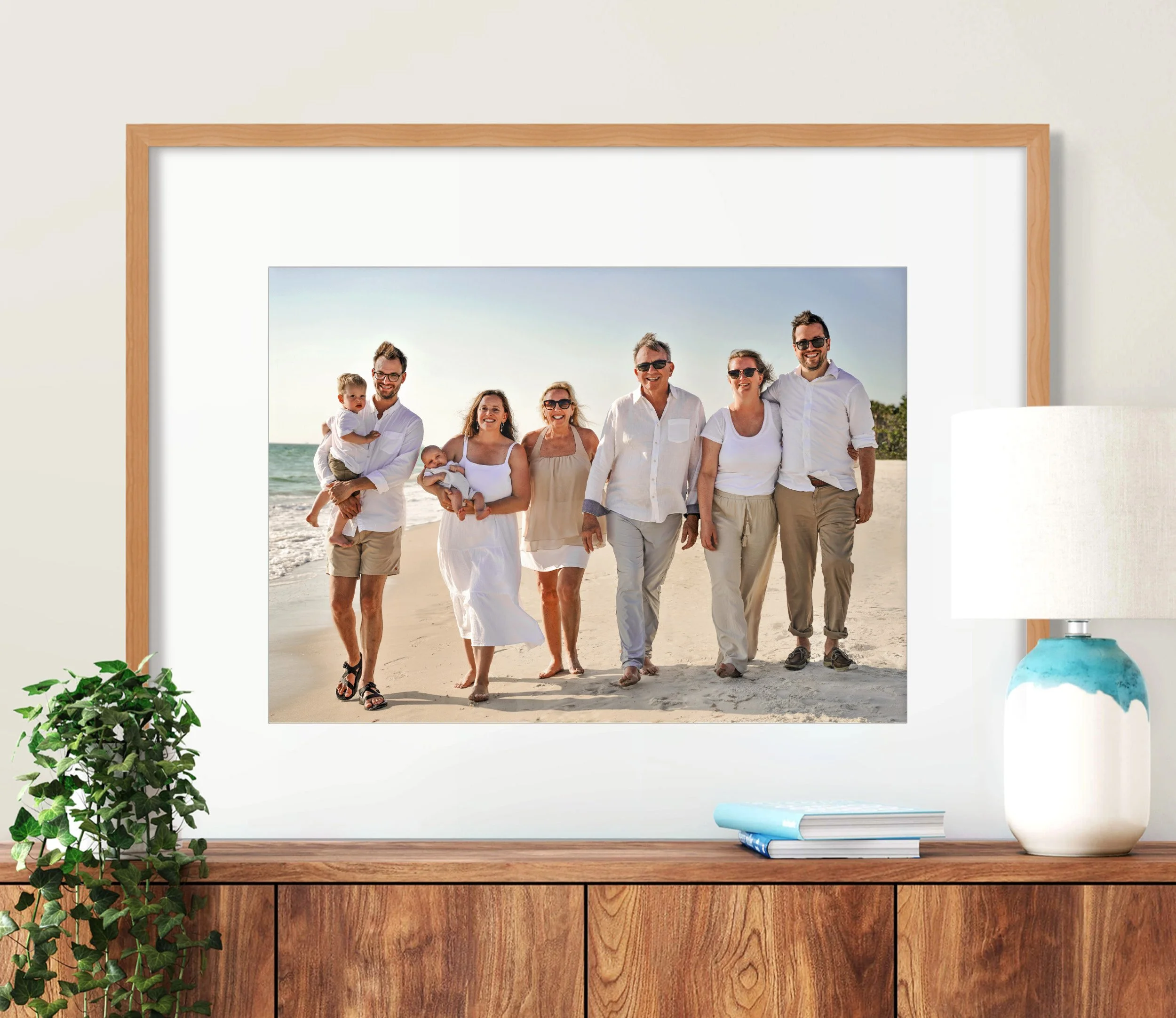 Family of seven walking on a beach in sunny weather, with a man holding a child and another woman holding a baby. The group is smiling and dressed in white and beige casual clothes.