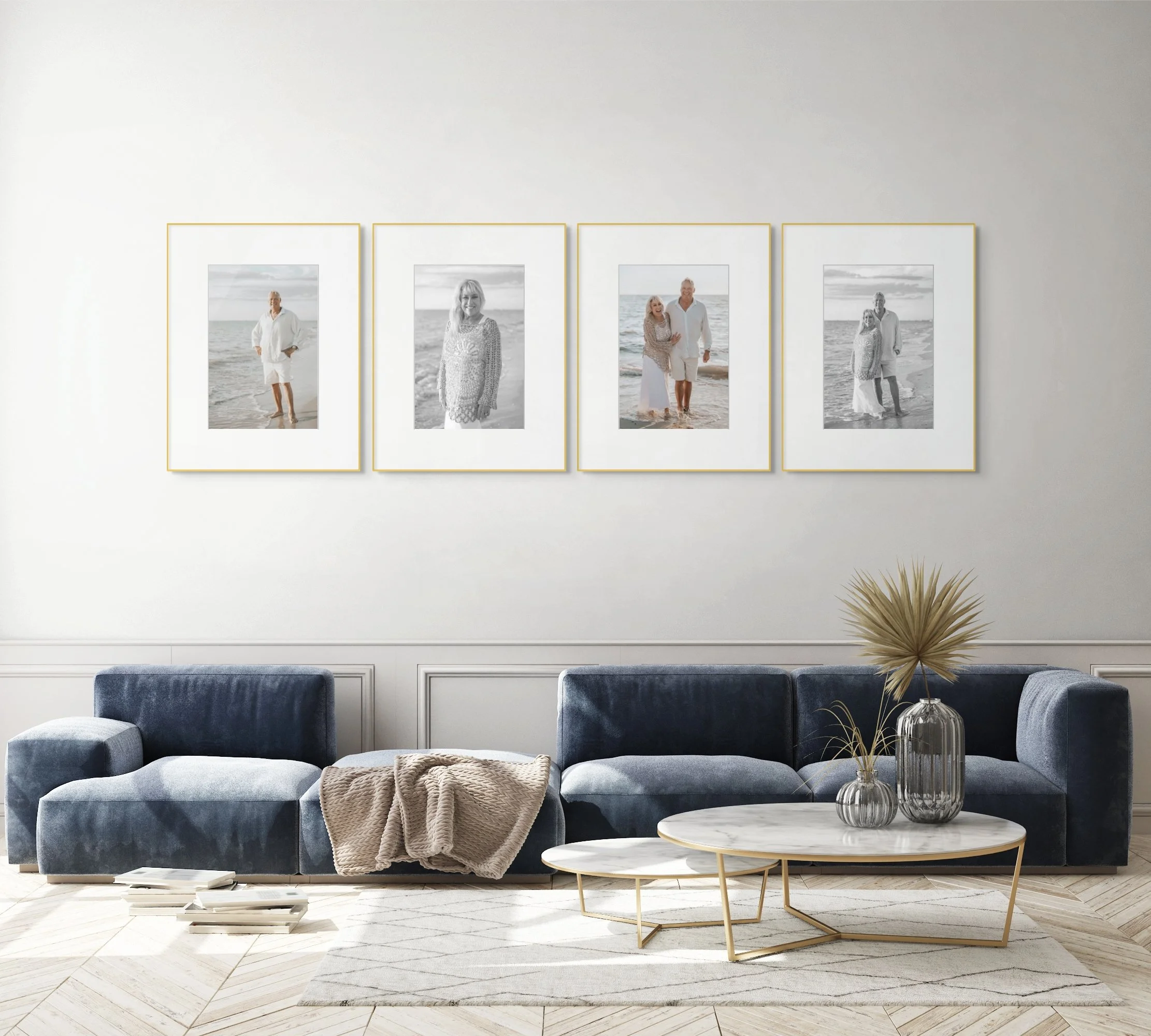 Living room with a dark blue sectional sofa, a beige throw blanket, two round nested marble-top tables with gold legs, and three decorative vases with dried plants. Four framed photos of elderly couple on the beach hang on the white wall behind.