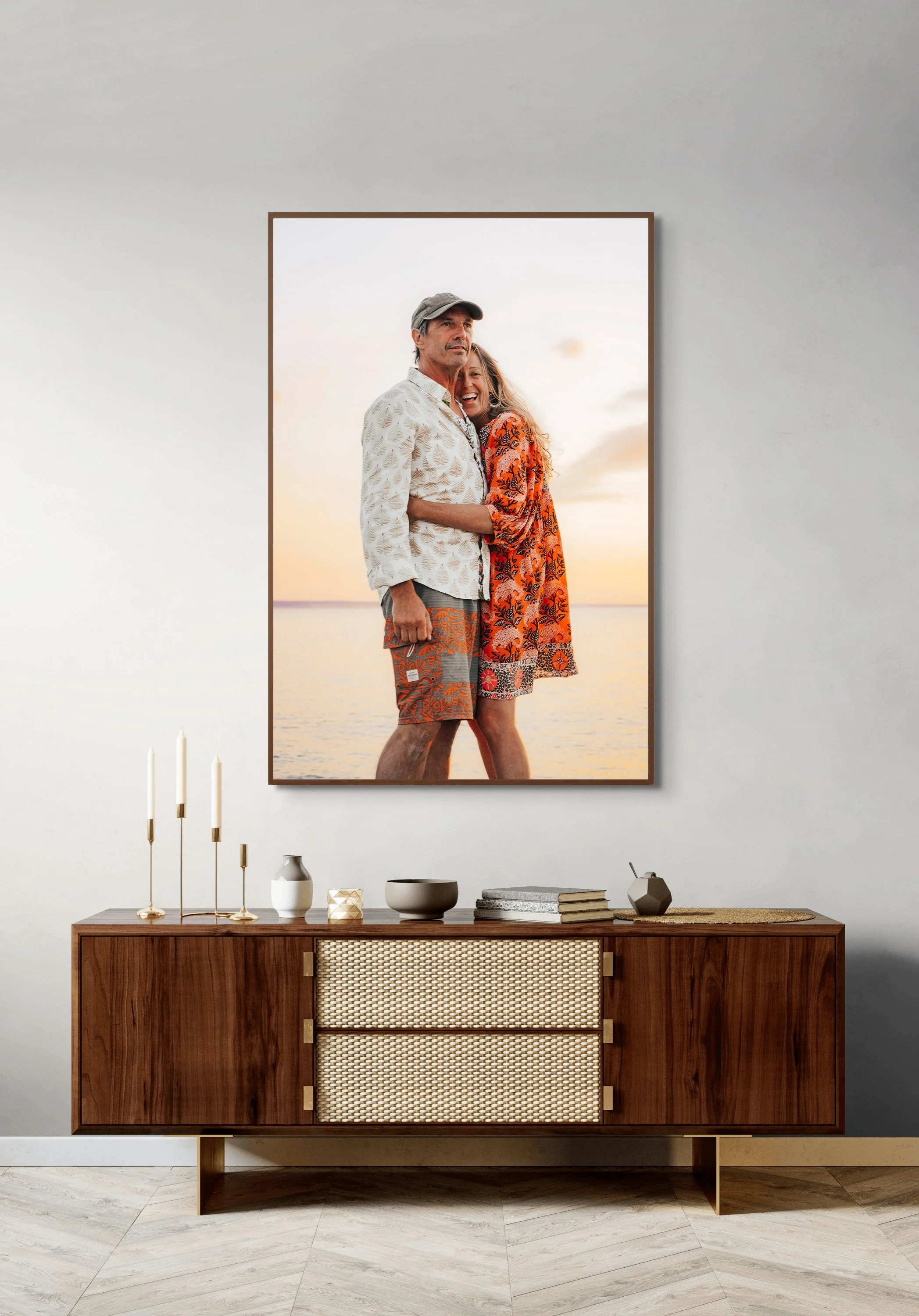 A large framed photograph of a man and woman at the beach, smiling at sunset, is hung on a featureless white wall above a wooden mid-century modern sideboard decorated with candles, vases, and books.