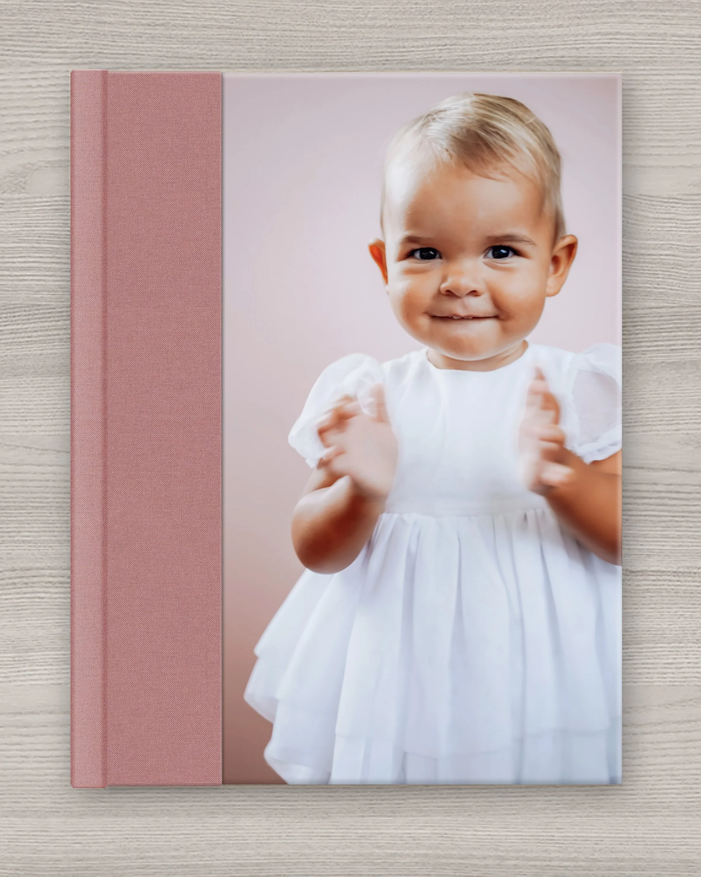 A pink hardcover photo book with a picture of a young girl with short blonde hair, wearing a white dress, smiling, and making a thumbs-up gesture in front of a pink background. The book is placed on a light-colored wooden surface.