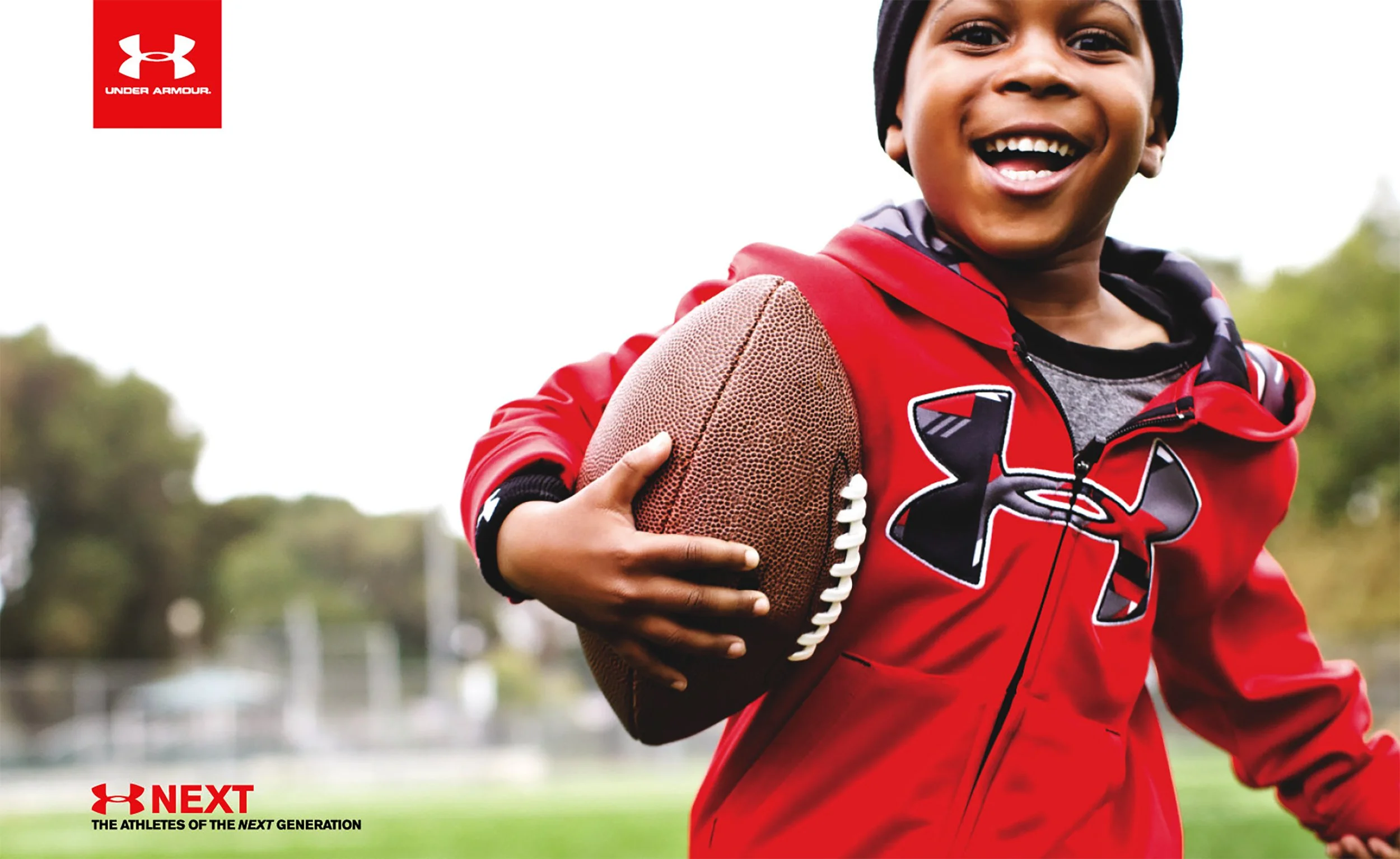 Young boy smiling and running outdoors, holding an American football, wearing a red Under Armour hoodie with black and white accents, on a sports field with a blurred background.