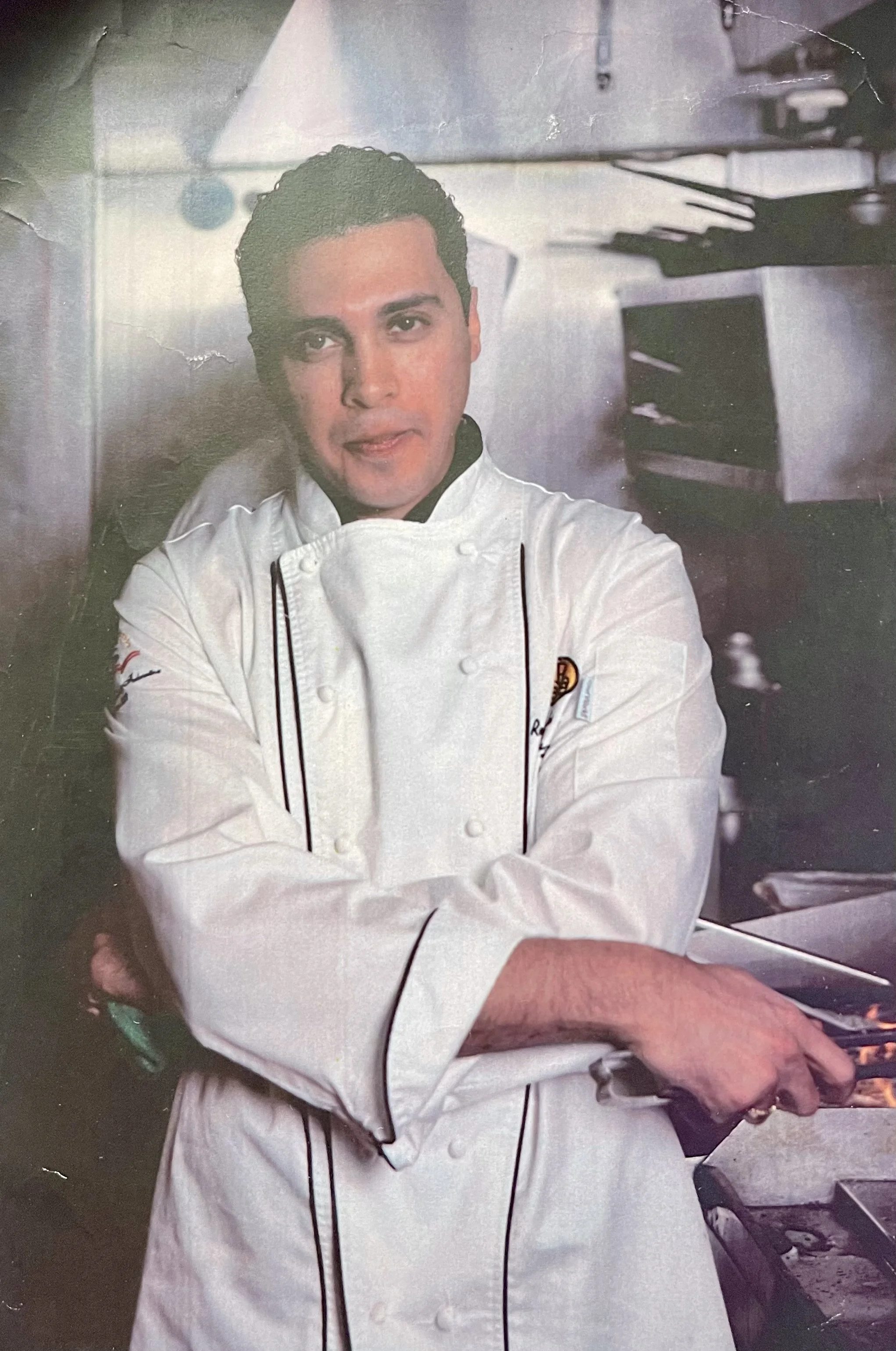 A male chef in a white chef's coat standing in a professional kitchen, holding a spatula and flipping food in a frying pan.