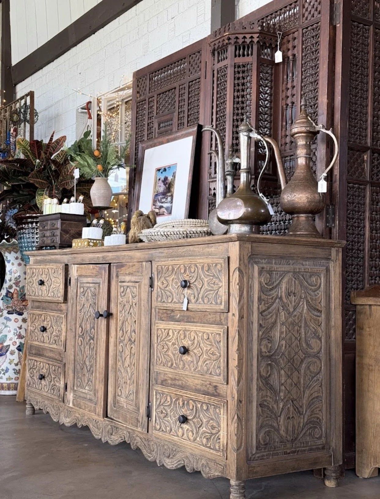 A rustic wooden sideboard with intricate carvings on the doors, set against a textured white brick wall. Decor items include vintage metal pitchers, a framed artwork, a potted plant, and various small accessories.