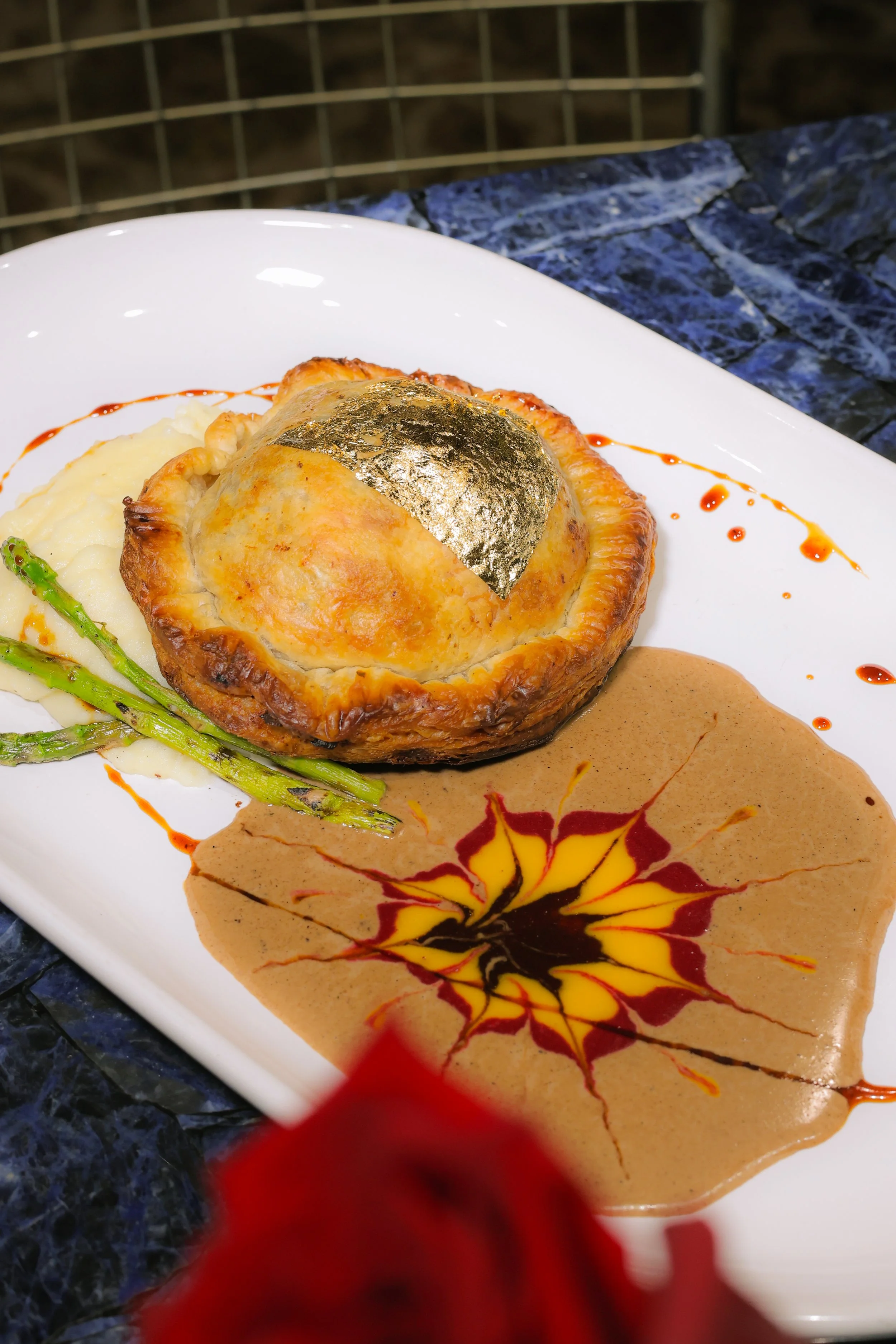 A plated dish featuring a baked pastry with a piece of fish on top, green asparagus, mashed potatoes, and a decorative sauce with a colorful pattern.