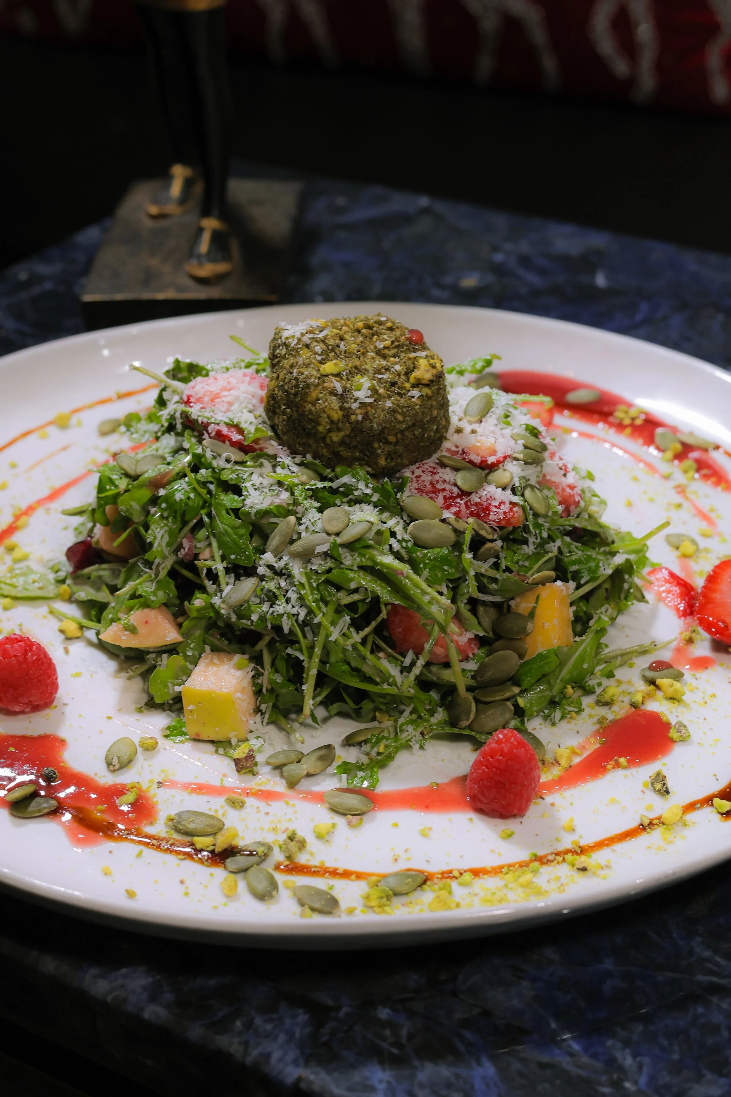 A colorful mixed green salad with strawberries, diced cheese, pumpkin seeds, drizzled with dressing, and topped with a round fried or croquette food item on a white plate.