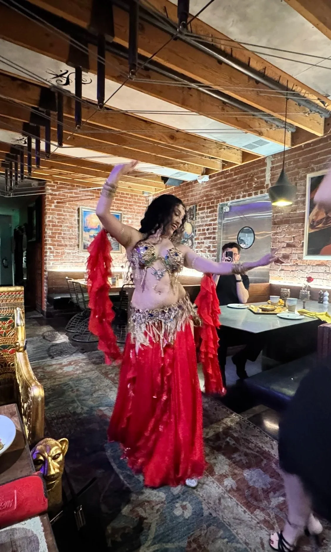 A woman performing a belly dance in a restaurant with exposed brick walls, seated guests, and decorative paintings.