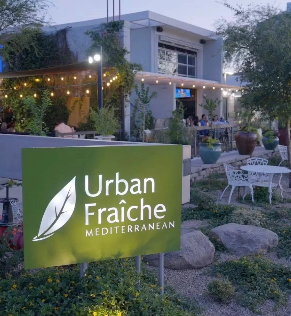 Outdoor seating area of a restaurant called Urban Fraîche Mediterranean with string lights, tables, chairs, and plants, in the evening.