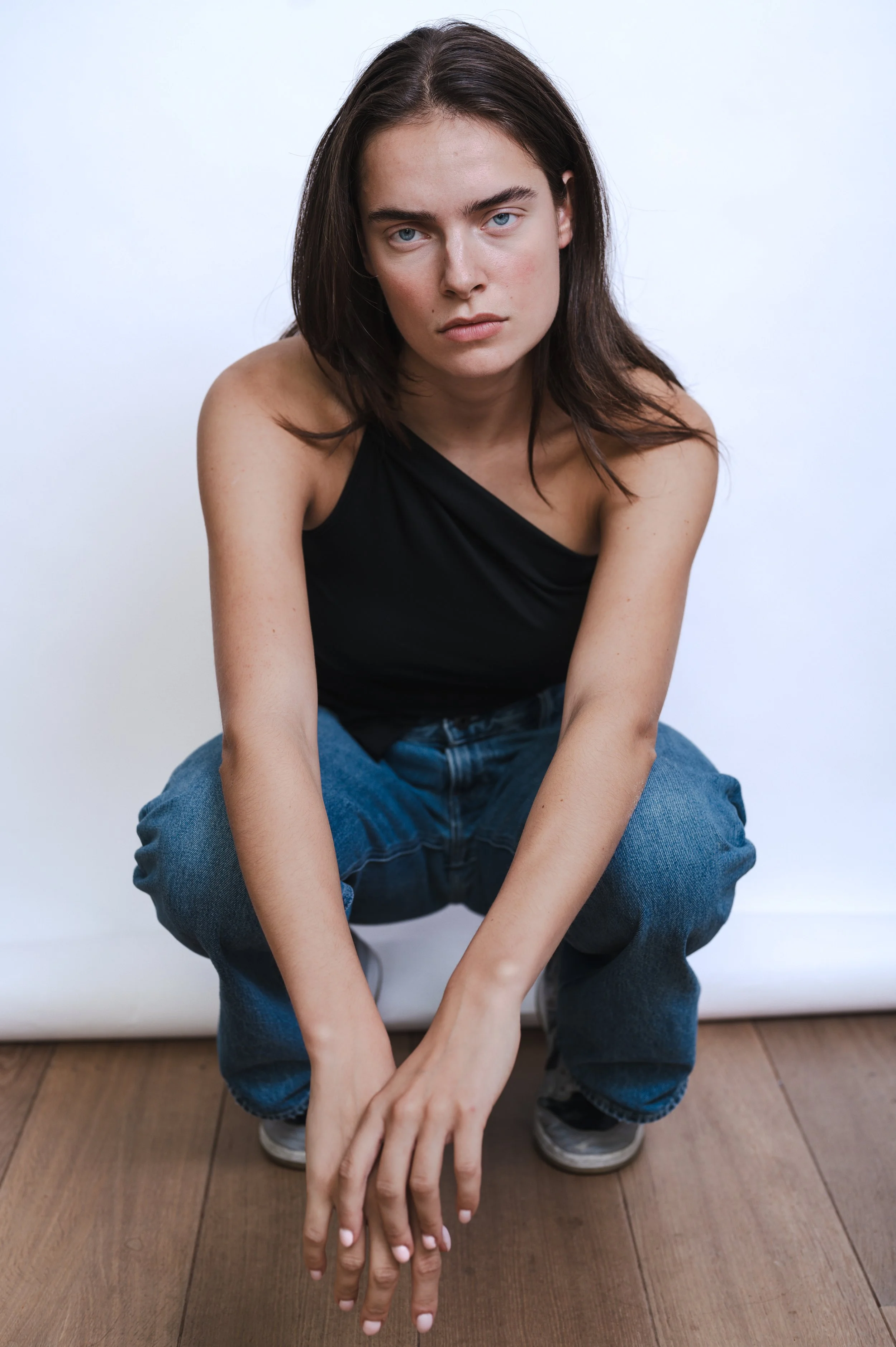 A young woman with dark hair and blue eyes crouching on a wooden floor against a white background, wearing a black sleeveless top and blue jeans.