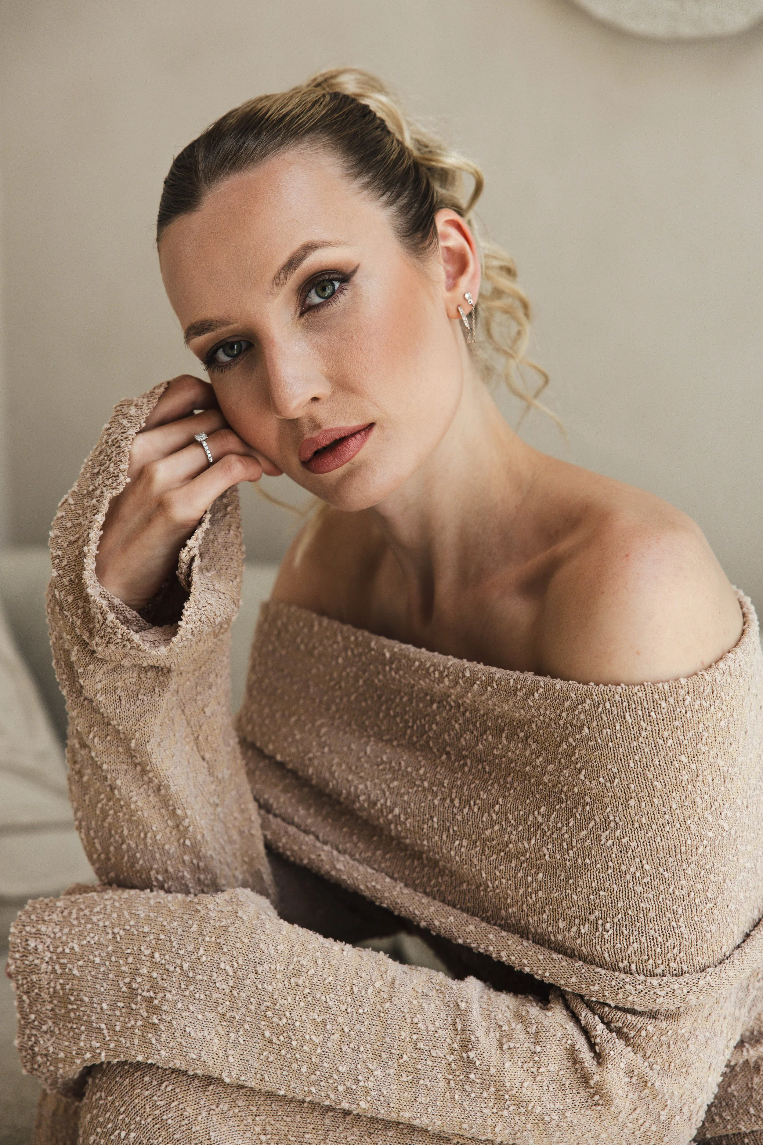 A woman with blonde hair pulled back, wearing a nude-colored off-the-shoulder sweater, looking at the camera with her hand near her face, showcasing earrings and a ring.