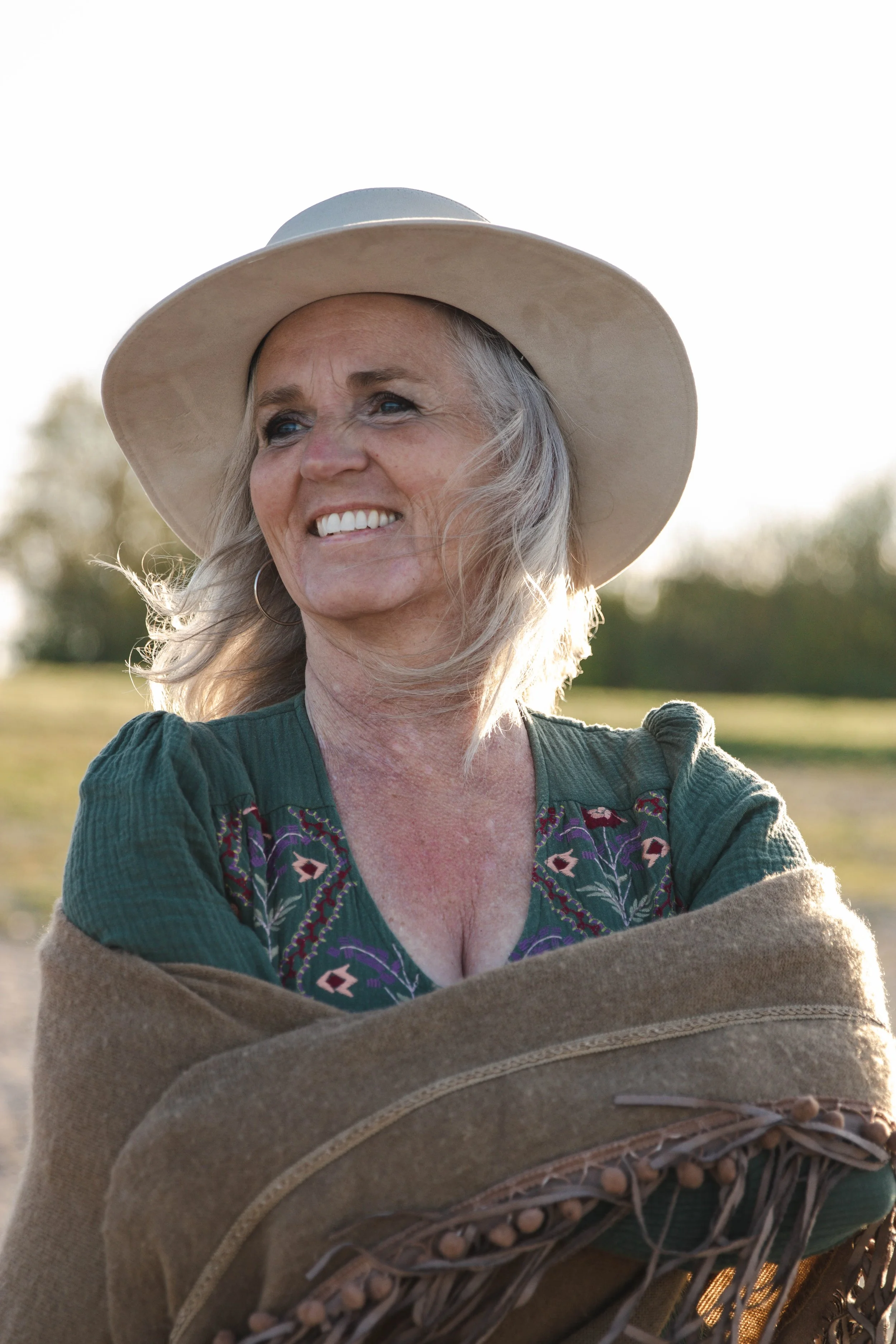 A woman smiling outside during golden hour, wearing a large hat and a green embroidered top, with a warm, peaceful expression.