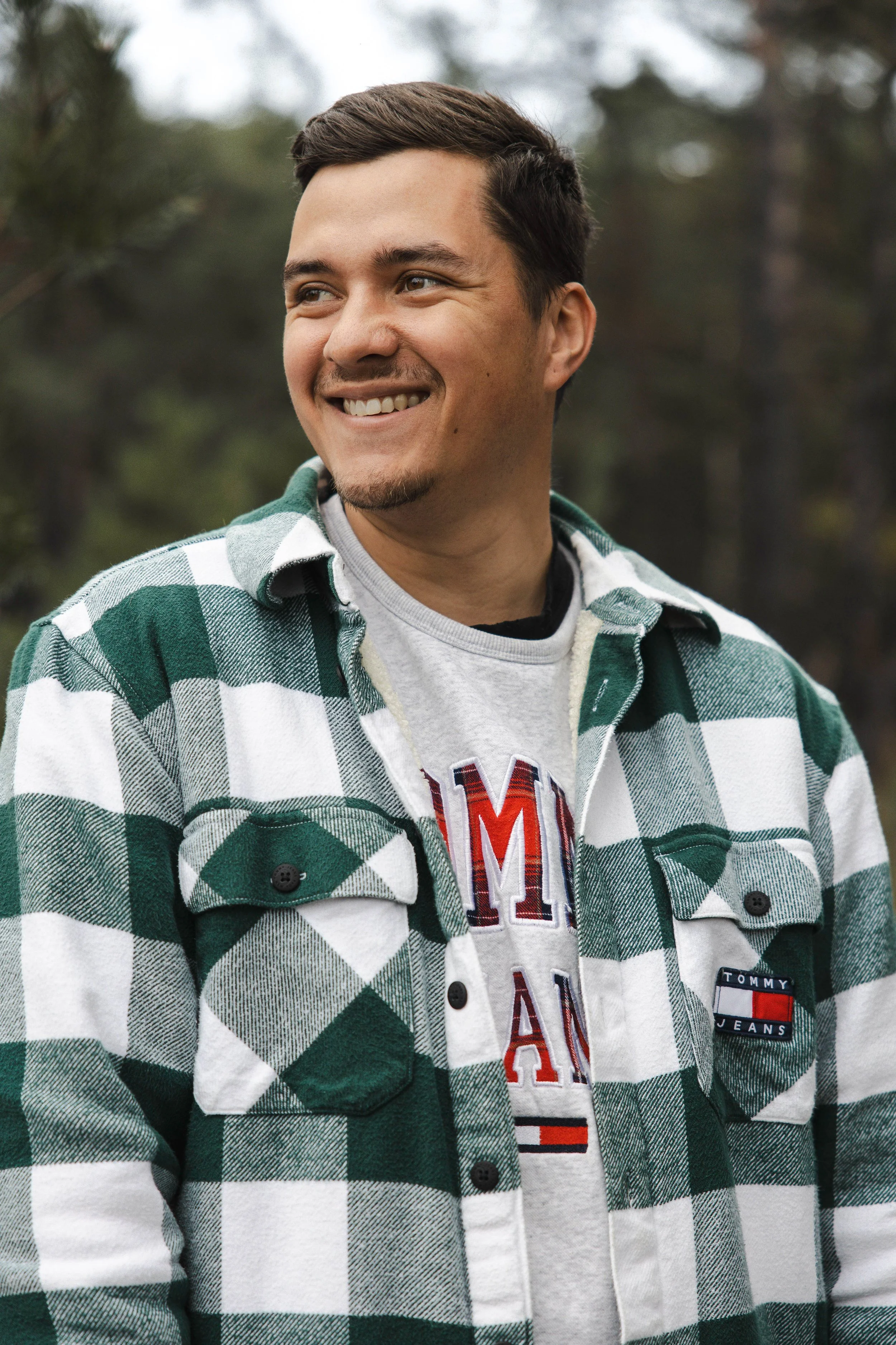 A young man with short dark hair and a goatee, smiling and looking to the side, outdoors with trees in the background, wearing a green and white plaid shirt over a gray graphic t-shirt.