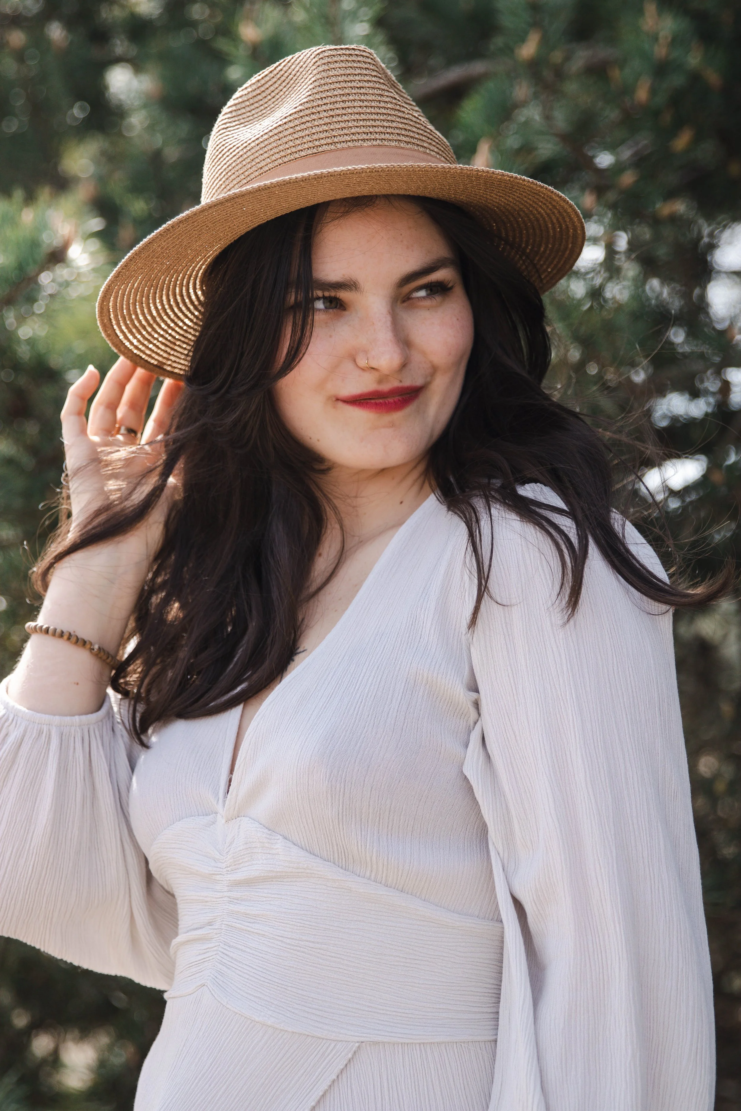 A woman with dark hair wearing a tan sunhat and white dress outdoors, smiling slightly with trees in the background.