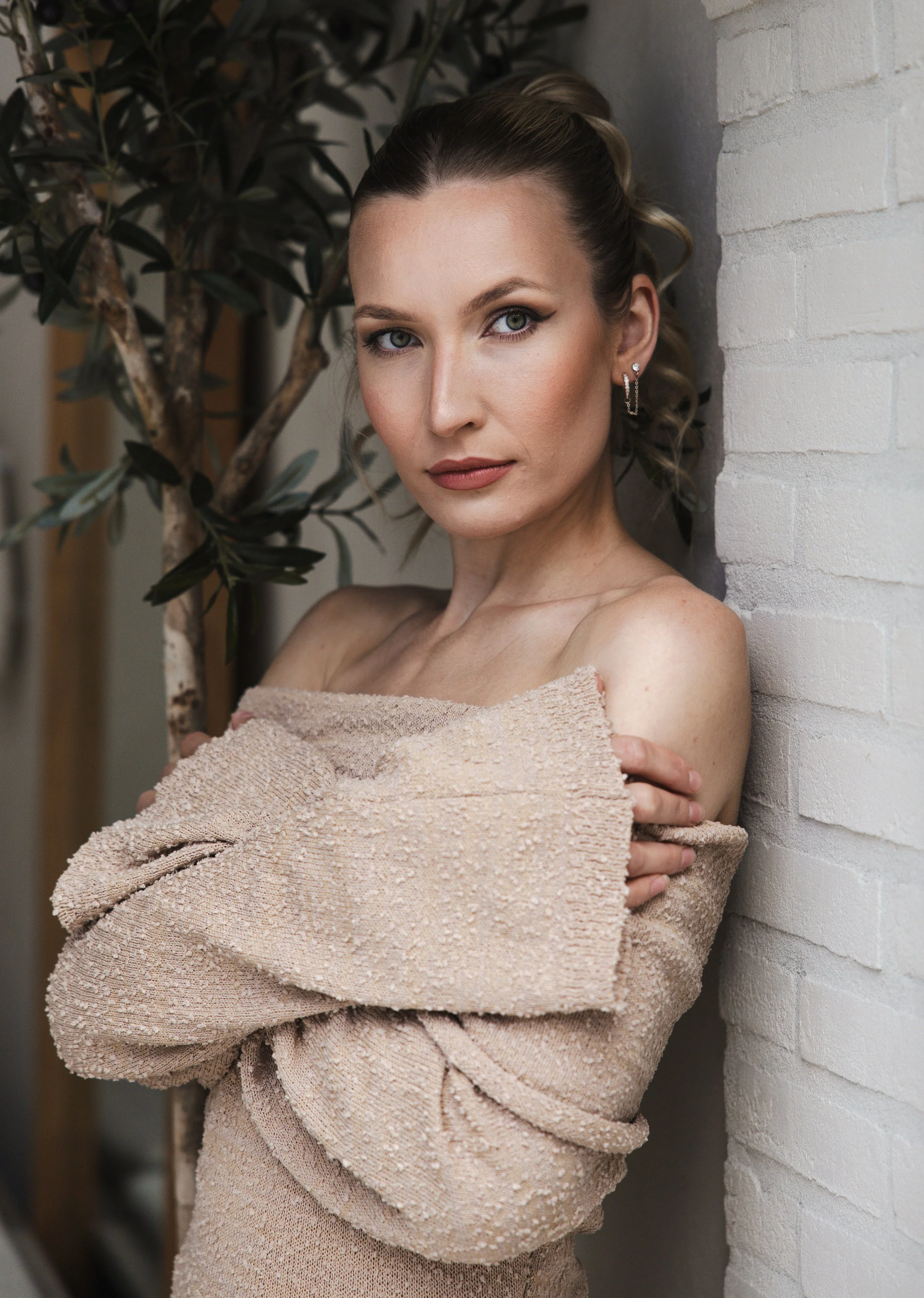 A woman with blonde hair styled in loose curls, wearing earrings, and wrapped in a textured beige sweater, stands against a white brick wall near a potted plant, looking directly at the camera.