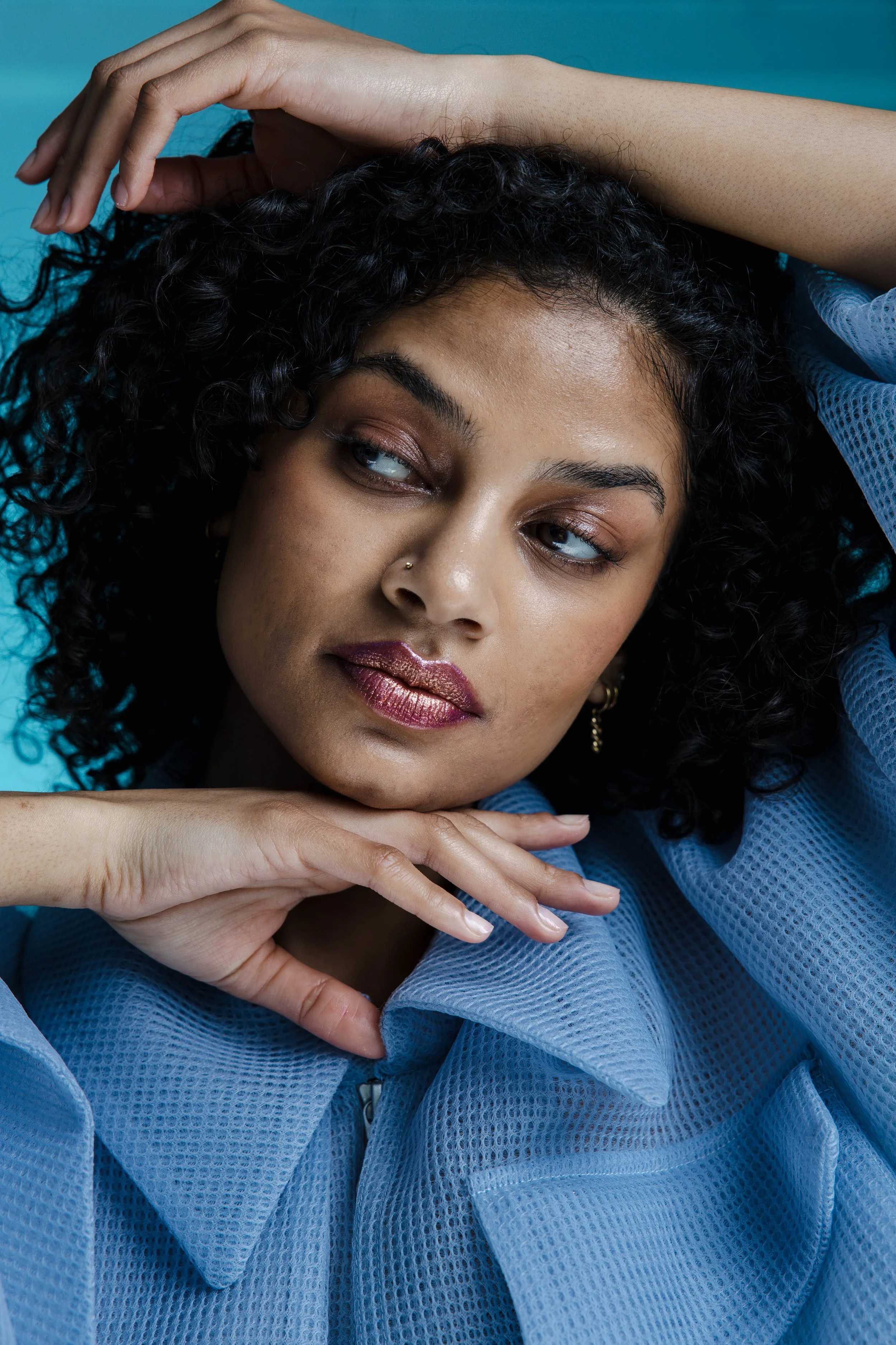 A woman with curly black hair, light brown skin, and pinkish metallic lipstick, wearing a blue mesh jacket, poses with her right hand resting on her forehead and her left hand under her chin against a blue background.