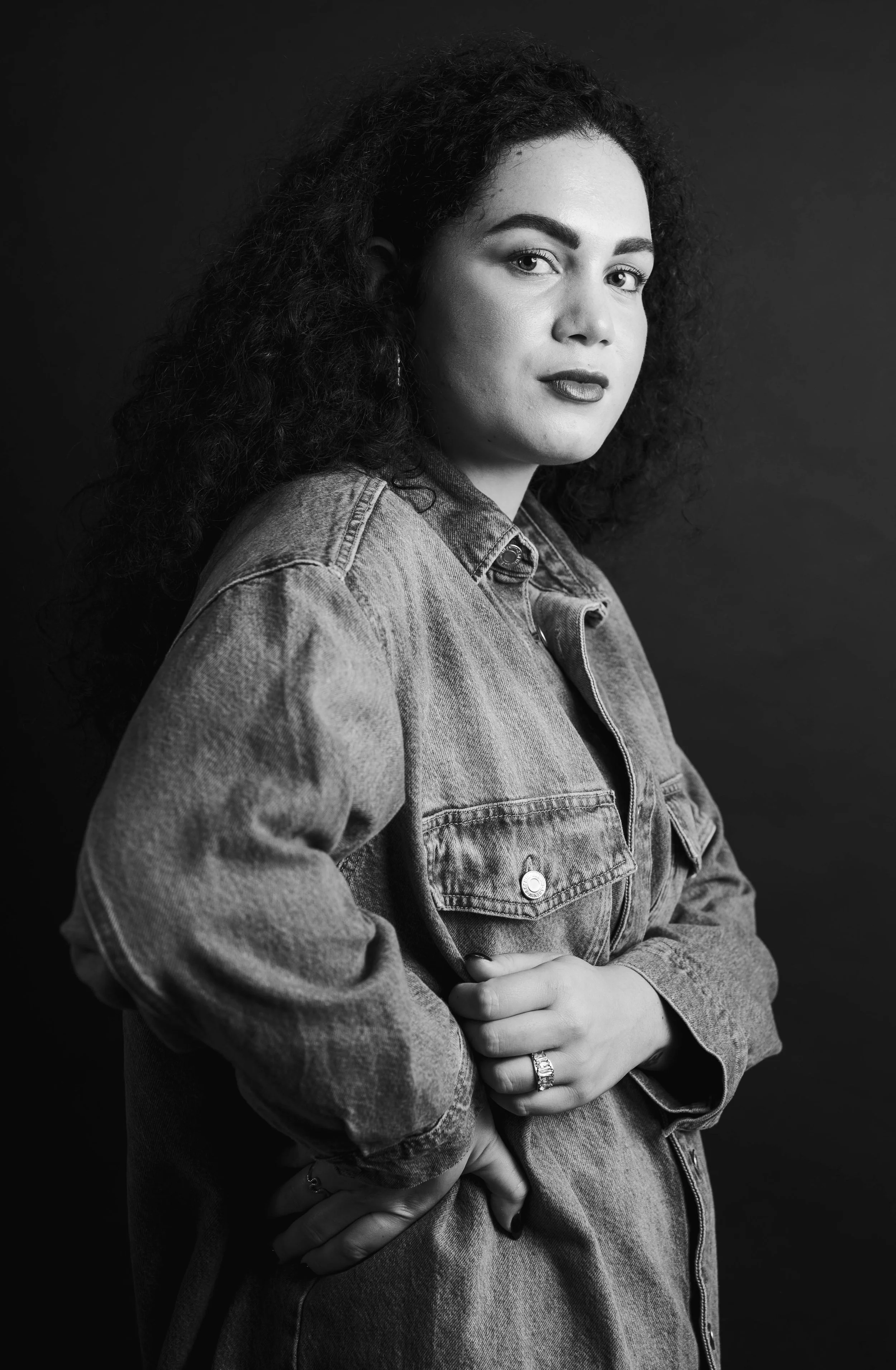 Black and white portrait of a woman with curly hair, wearing a denim jacket, standing against a dark background.
