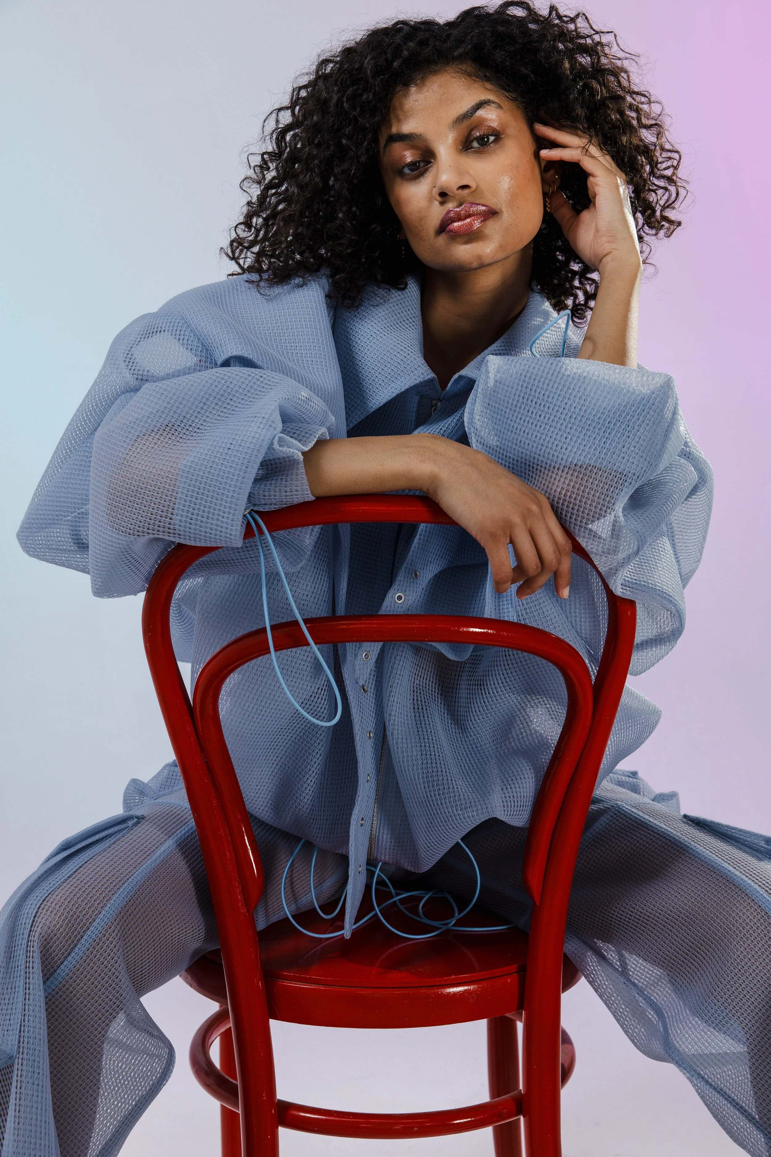 A woman with curly hair, dressed in a light blue oversized mesh shirt and matching pants, sitting on a red chair against a soft pastel background.