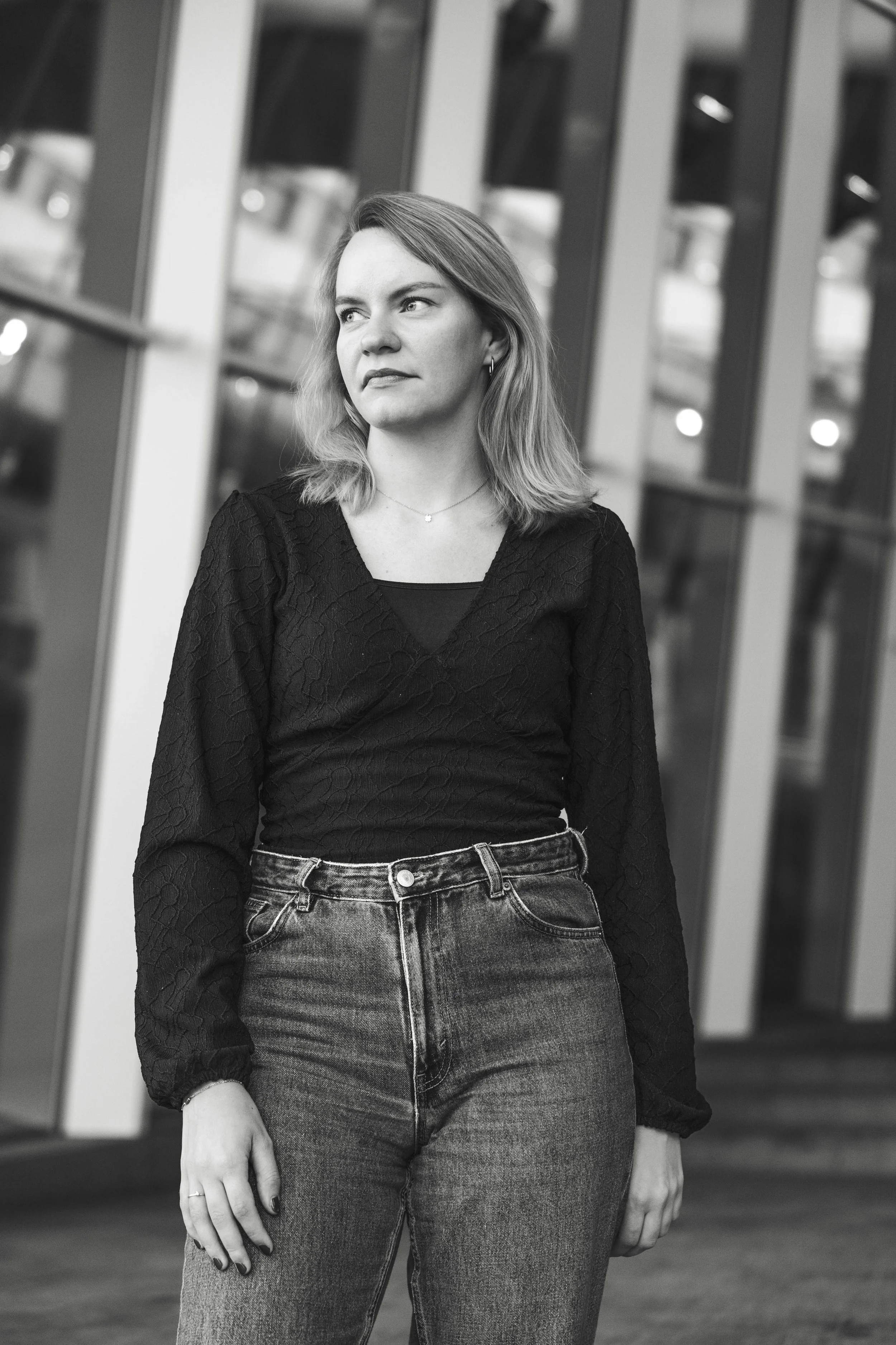 Black-and-white photo of a woman standing outdoors, looking to her left with a serious expression, in front of a glass building with reflective windows.