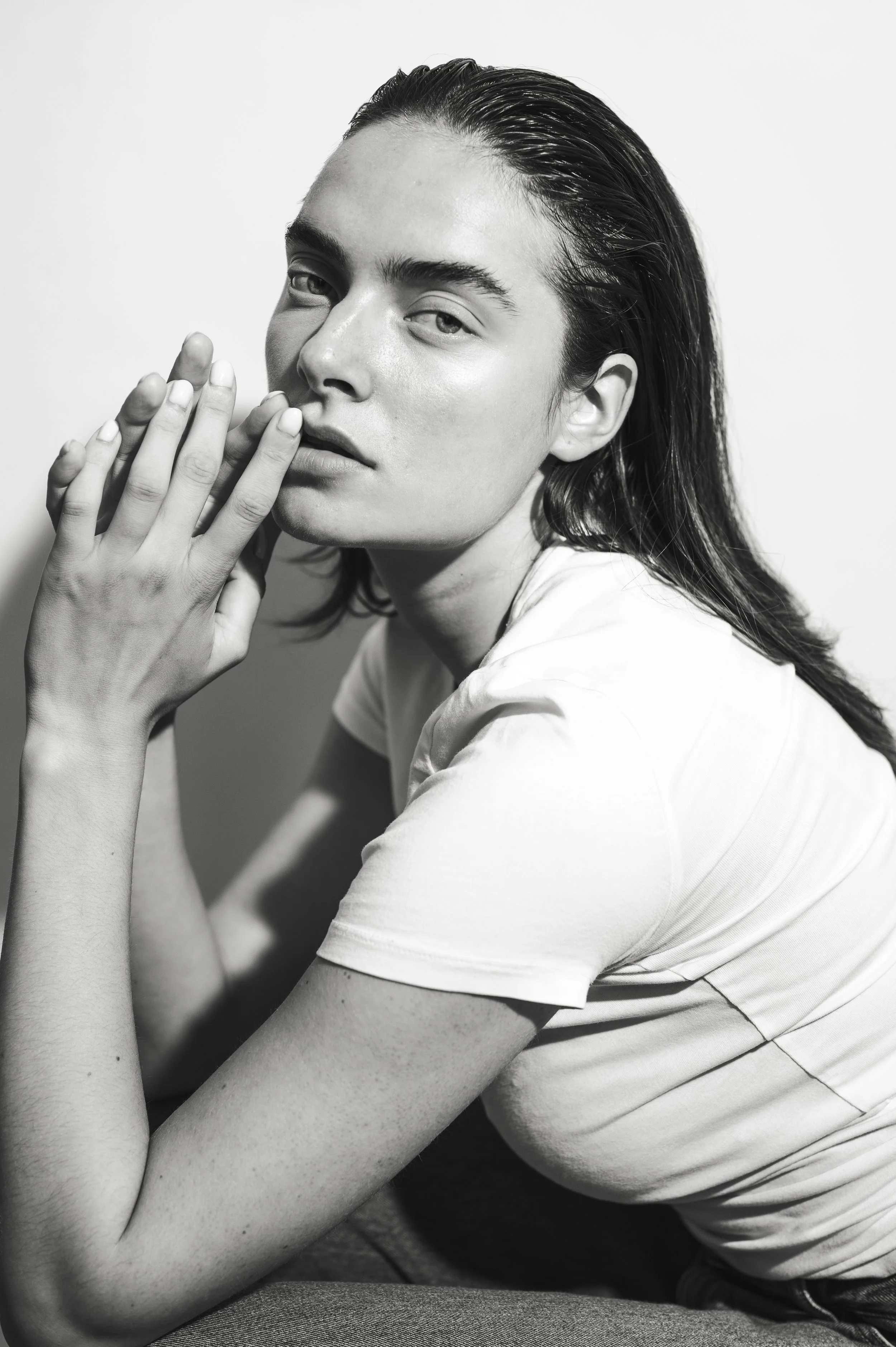 Black and white portrait of a young woman with wet hair, wearing a white t-shirt, looking at the camera with a relaxed expression, touching her face with her right hand.