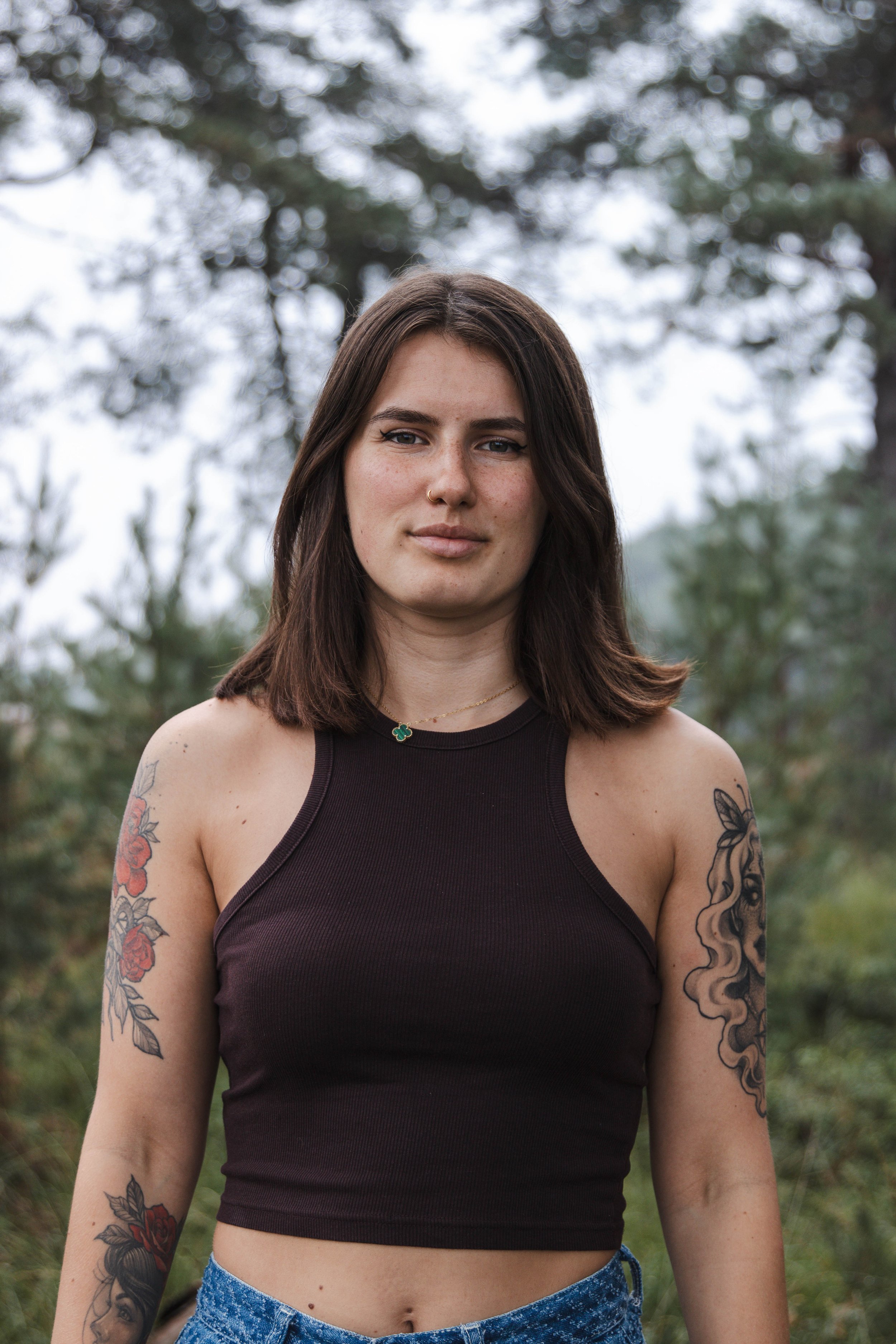 A young woman with shoulder-length brown hair, wearing a black sleeveless top, standing outdoors with trees in the background.