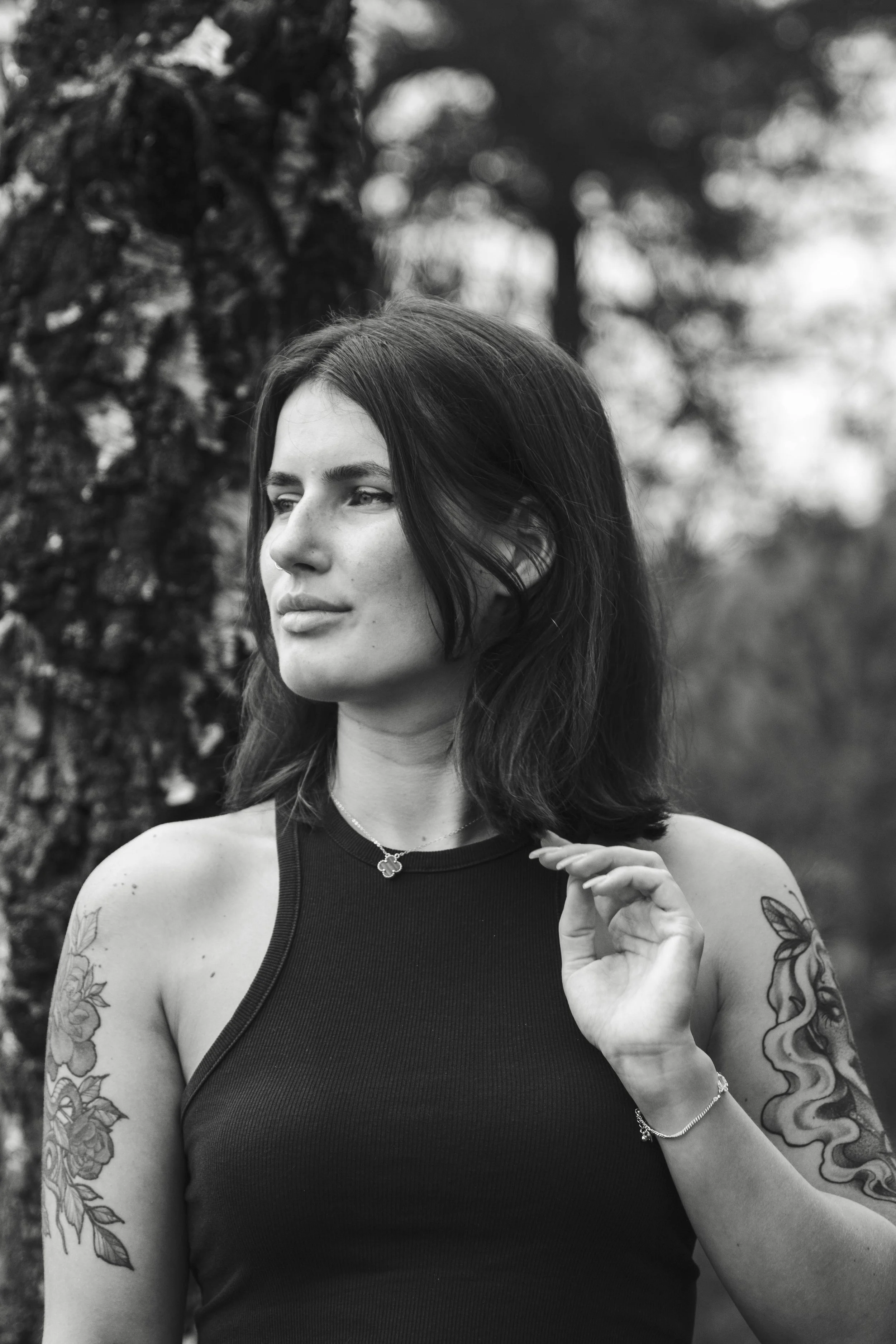 Black and white photo of a young woman with dark hair, tattoos on her arms, wearing a sleeveless top, standing outdoors near a tree.