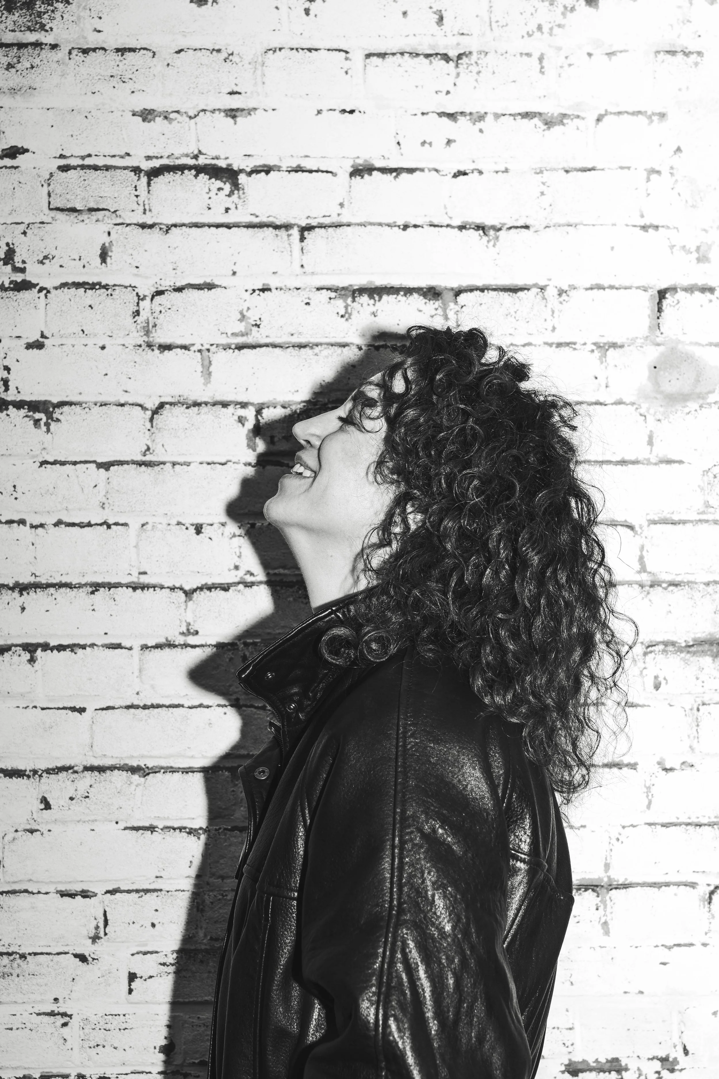 Black and white photo of a woman with curly hair wearing a leather jacket, smiling with her eyes closed, standing against a brick wall.