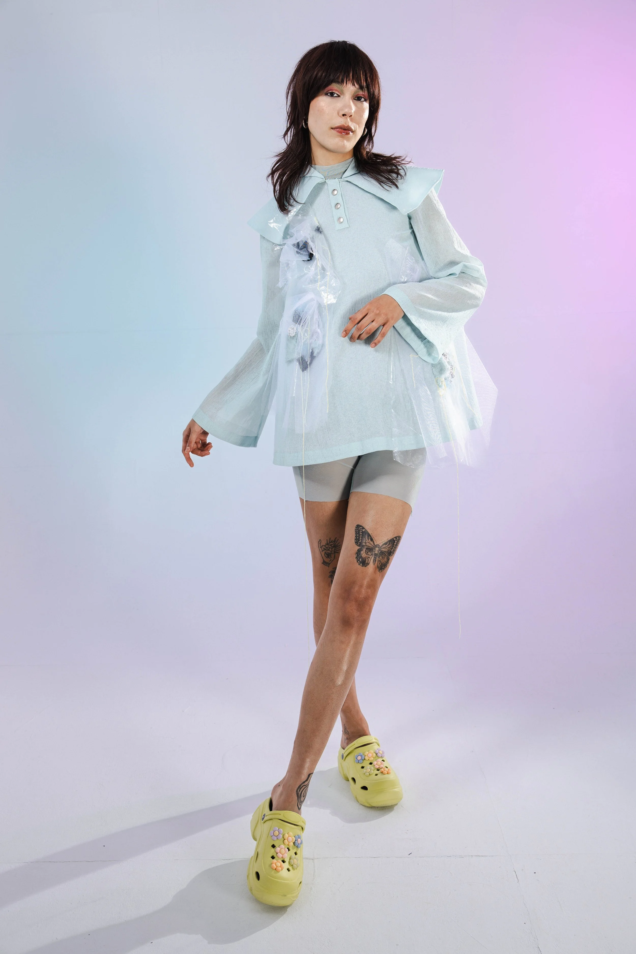 A woman with dark brown hair and tattoos on her thighs is standing in a studio with pastel pink and purple lighting. She is wearing a light blue oversized jacket with tulle details and yellow Crocs decorated with colorful flowers.