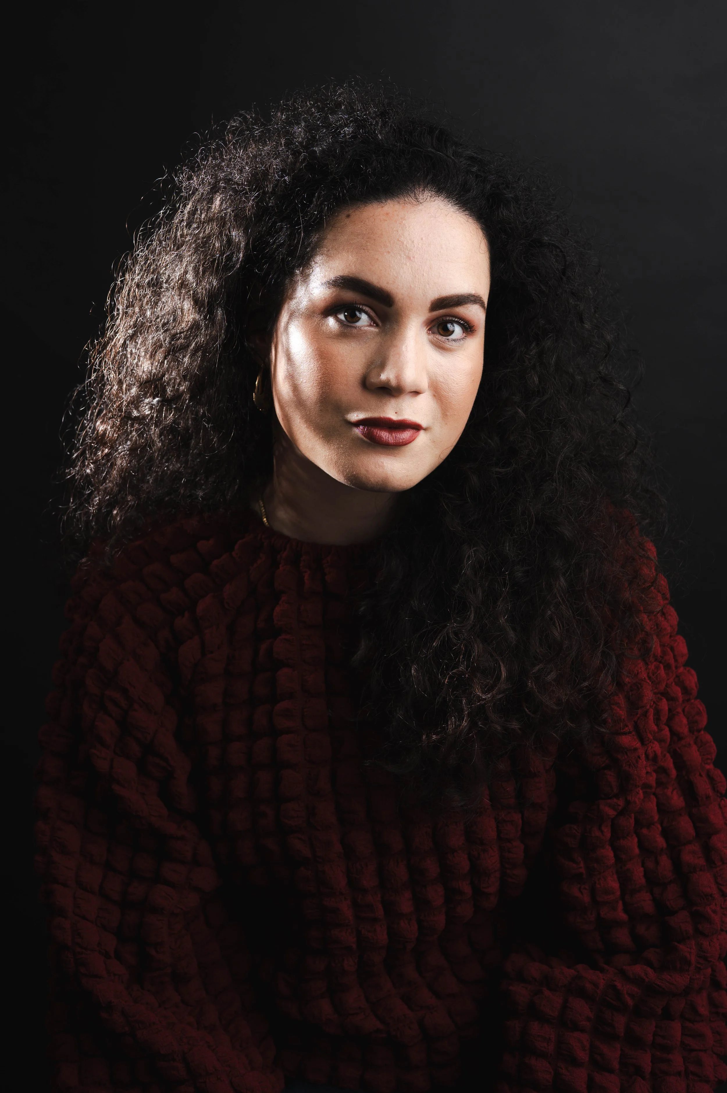Portrait of a woman with long, curly dark hair, wearing a textured burgundy sweater, against a dark background.
