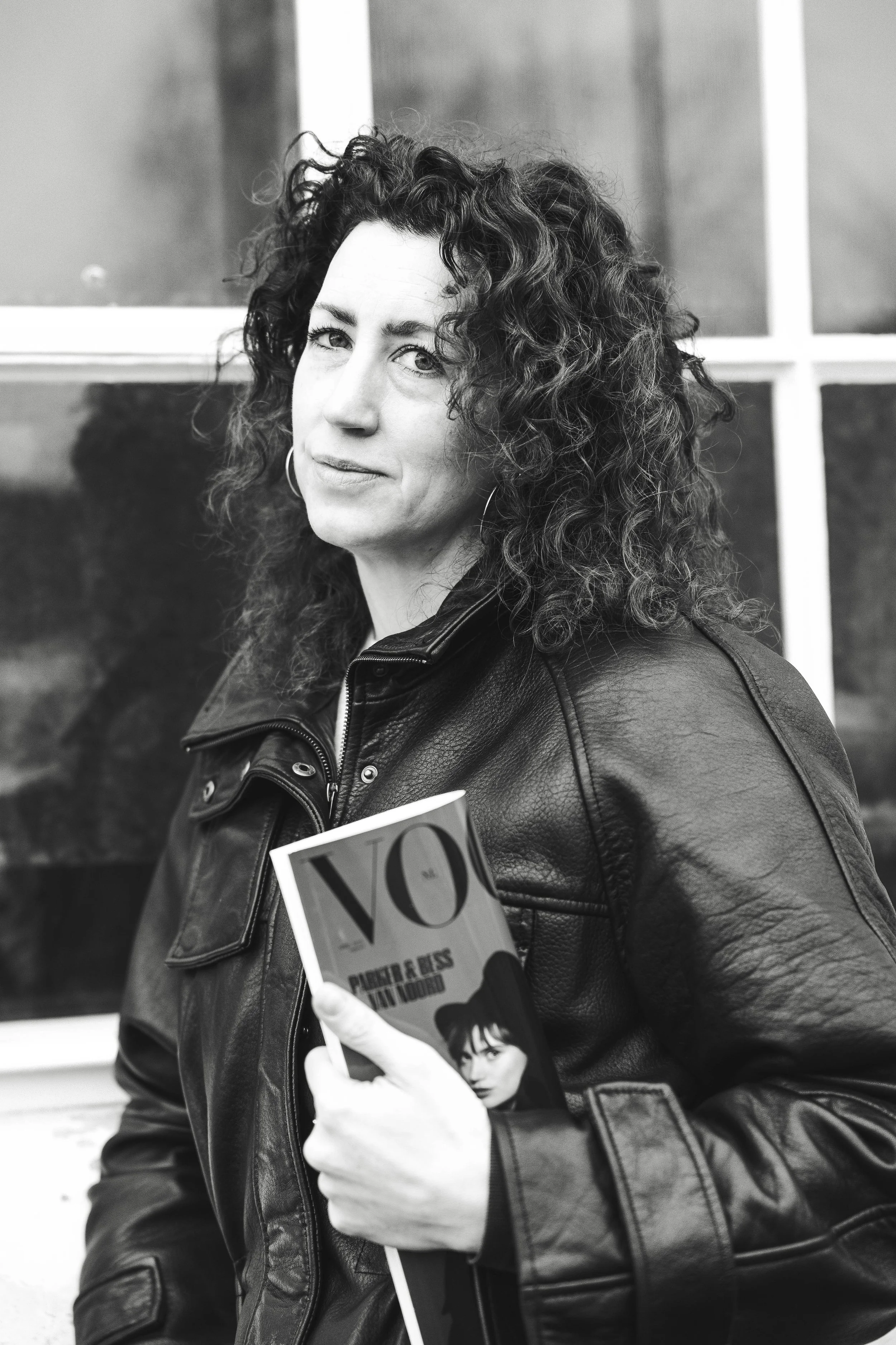 A woman with curly hair and hoop earrings wearing a leather jacket holding a magazine and standing in front of a window.