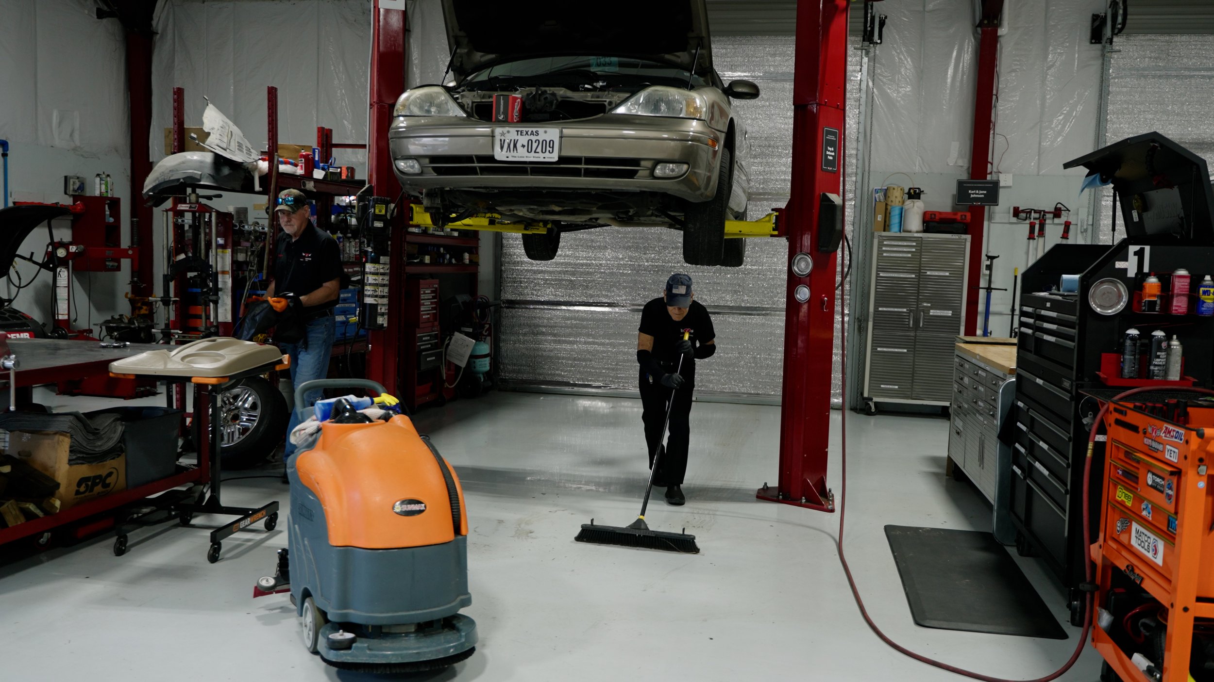 Automotive repair garage with a car on a lift, two mechanics working, one sweeping the floor, organized tools and equipment, shelves with supplies, and a vacuum cleaner.