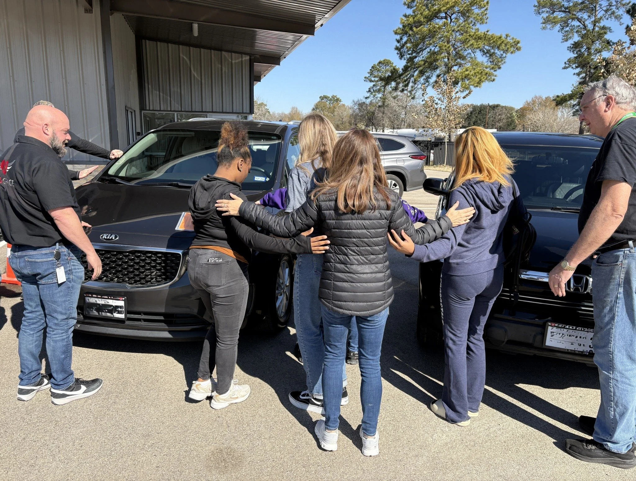 Group of people gathered in a circle, placing hands on each other's shoulders, near parked cars outside a building on a clear day.
