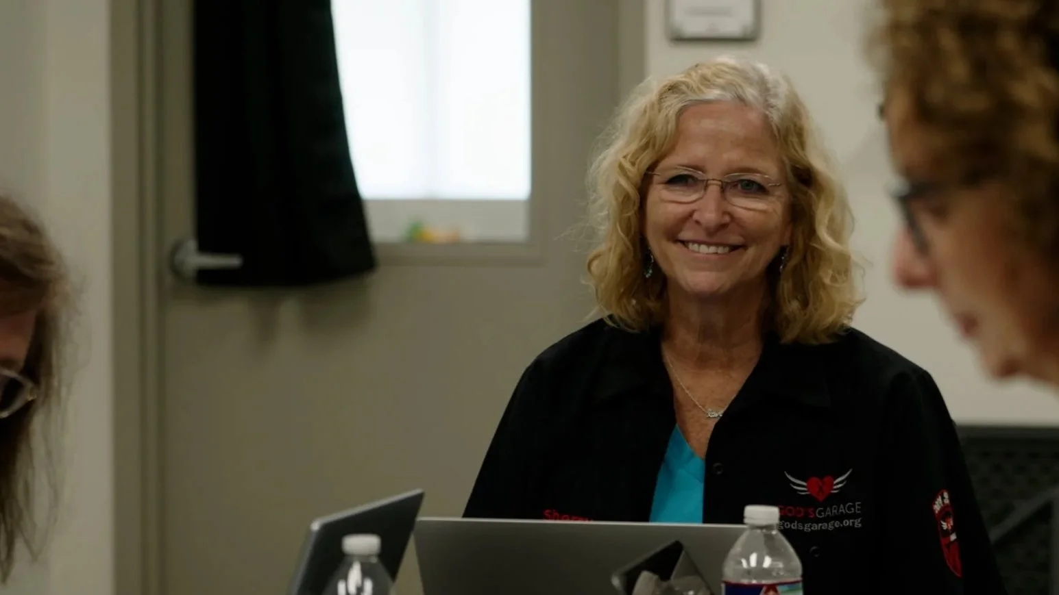 An older woman with gray hair and glasses holding and reading a piece of paper, standing with two other women in a room that appears to be an office or meeting room.