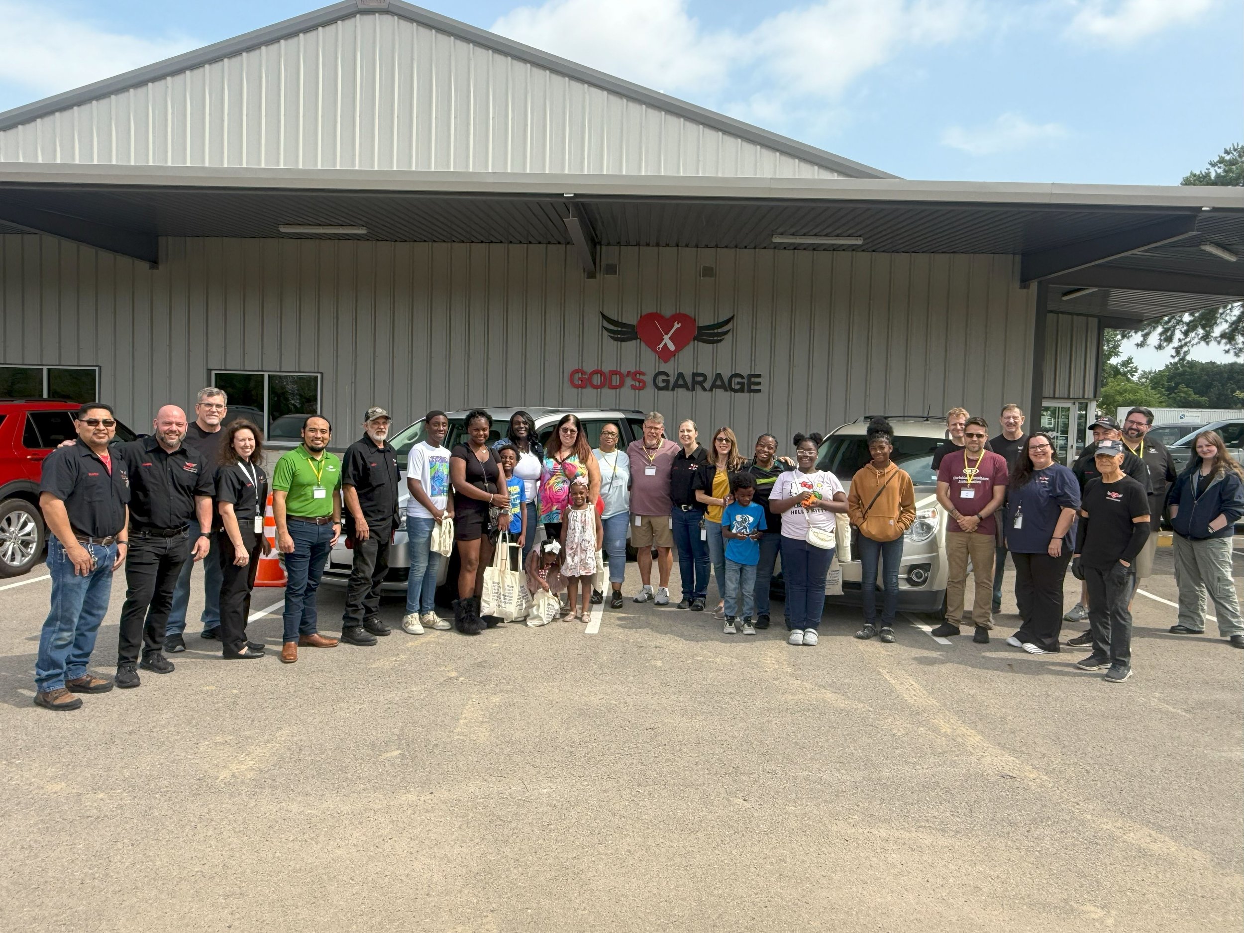 Group of people standing in front of a building that reads 'God's Garage' with a heart and wings logo, cars parked behind them, on a partly cloudy day.