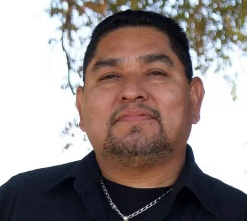 Close-up of a man with dark hair and facial hair, wearing a black shirt and silver chain, outdoors with trees in the background.