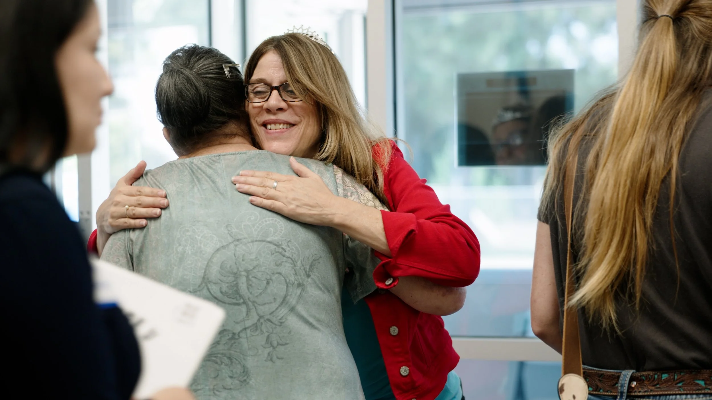 Two women hug in a warm embrace while standing near a large window, with other people nearby in an indoor setting.