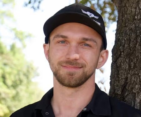 A young man with a beard and blue eyes standing outdoors next to a tree, wearing a black baseball cap and a black polo shirt.