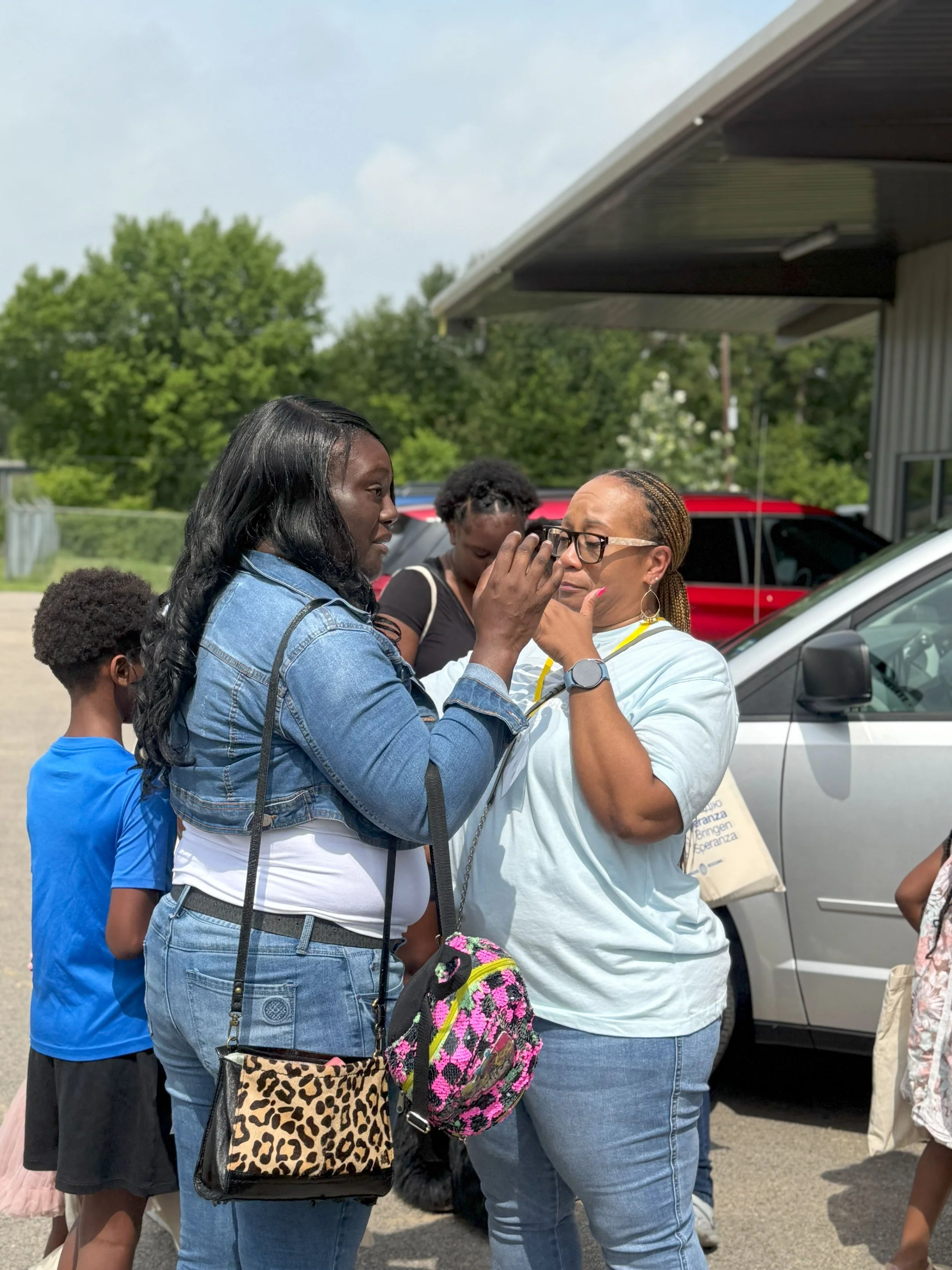 A woman in a denim jacket is comforting another woman, who is wiping her eyes, in an outdoor parking lot with children and vehicles around.