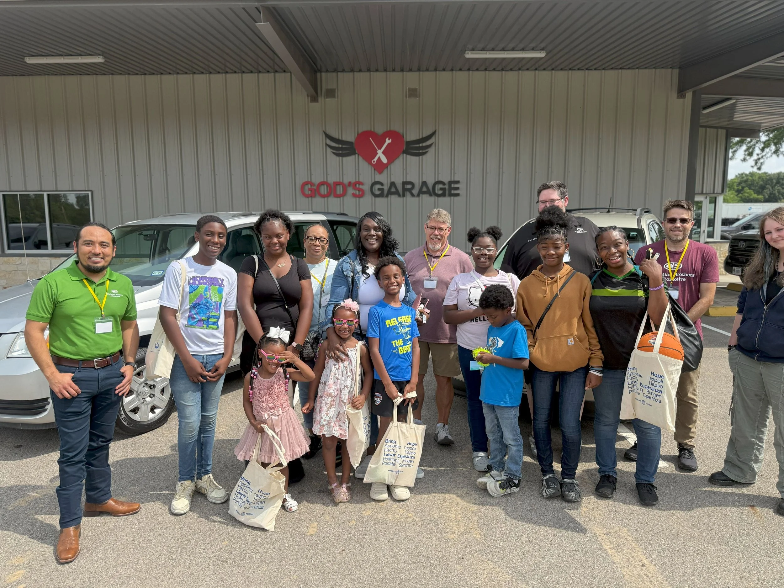 Group of people standing in front of a building with a sign that reads 'God's Garage' with a heart and wing logo, some holding bags and a basketball.