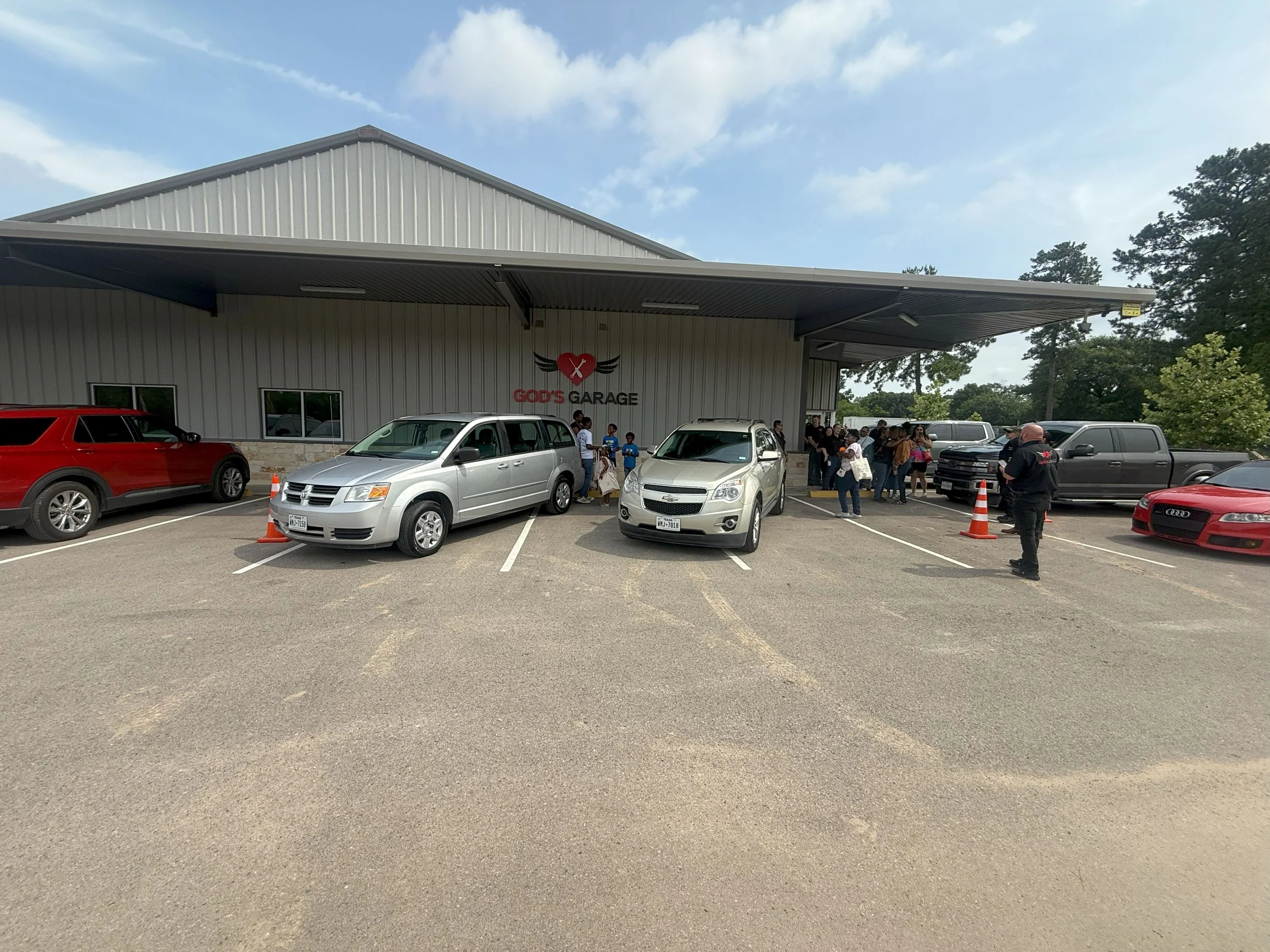 People gather outside a building labeled "God's Garage," with cars parked in front and some orange traffic cones around. The building has a metal roof and side walls, with a logo featuring a heart and wings.