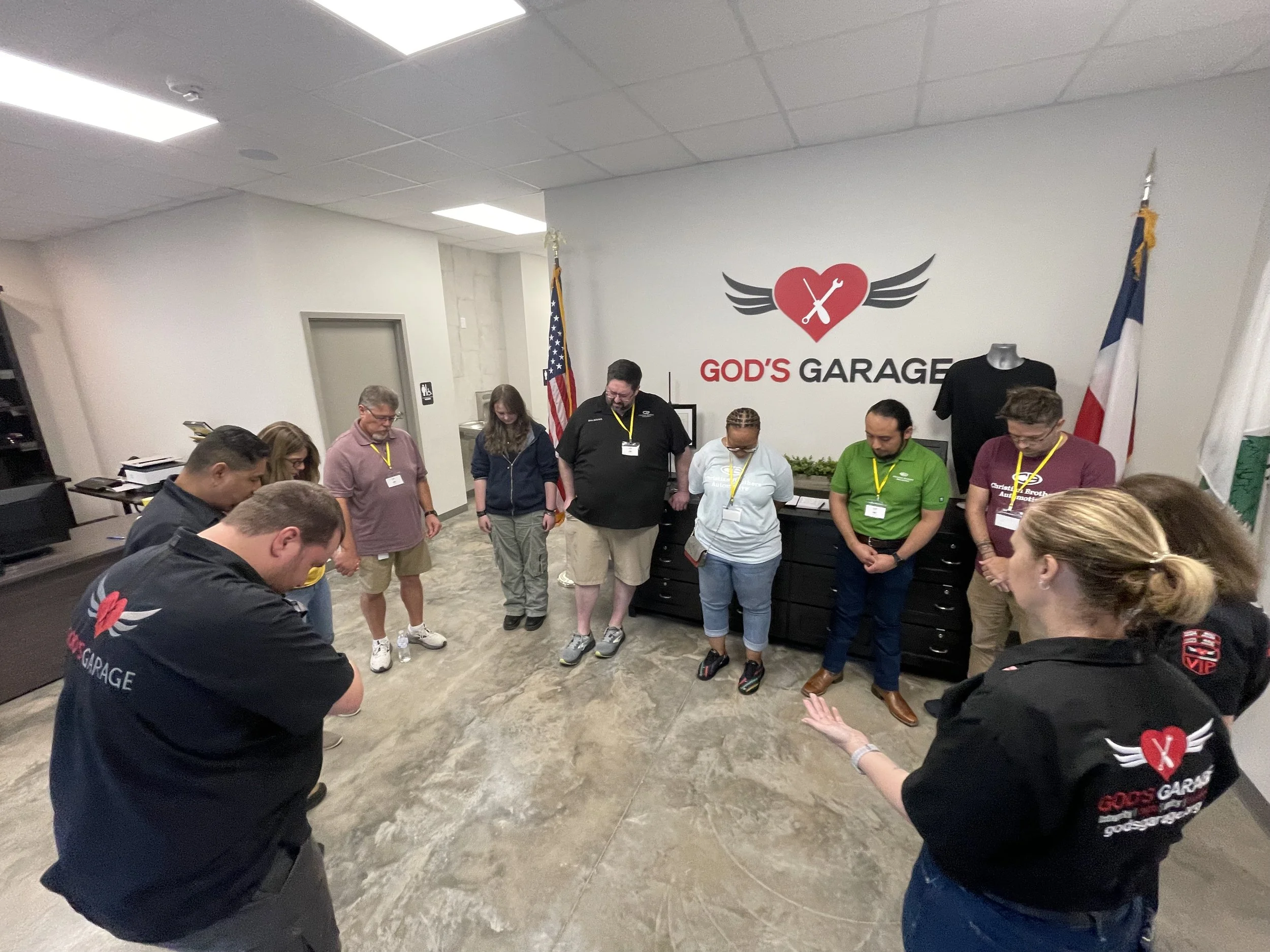A group of people standing in a circle in a room with a sign that reads 'GOD'S GARAGE' on the wall behind them, with some individuals wearing matching shirts with the same logo and others with yellow lanyards around their necks.