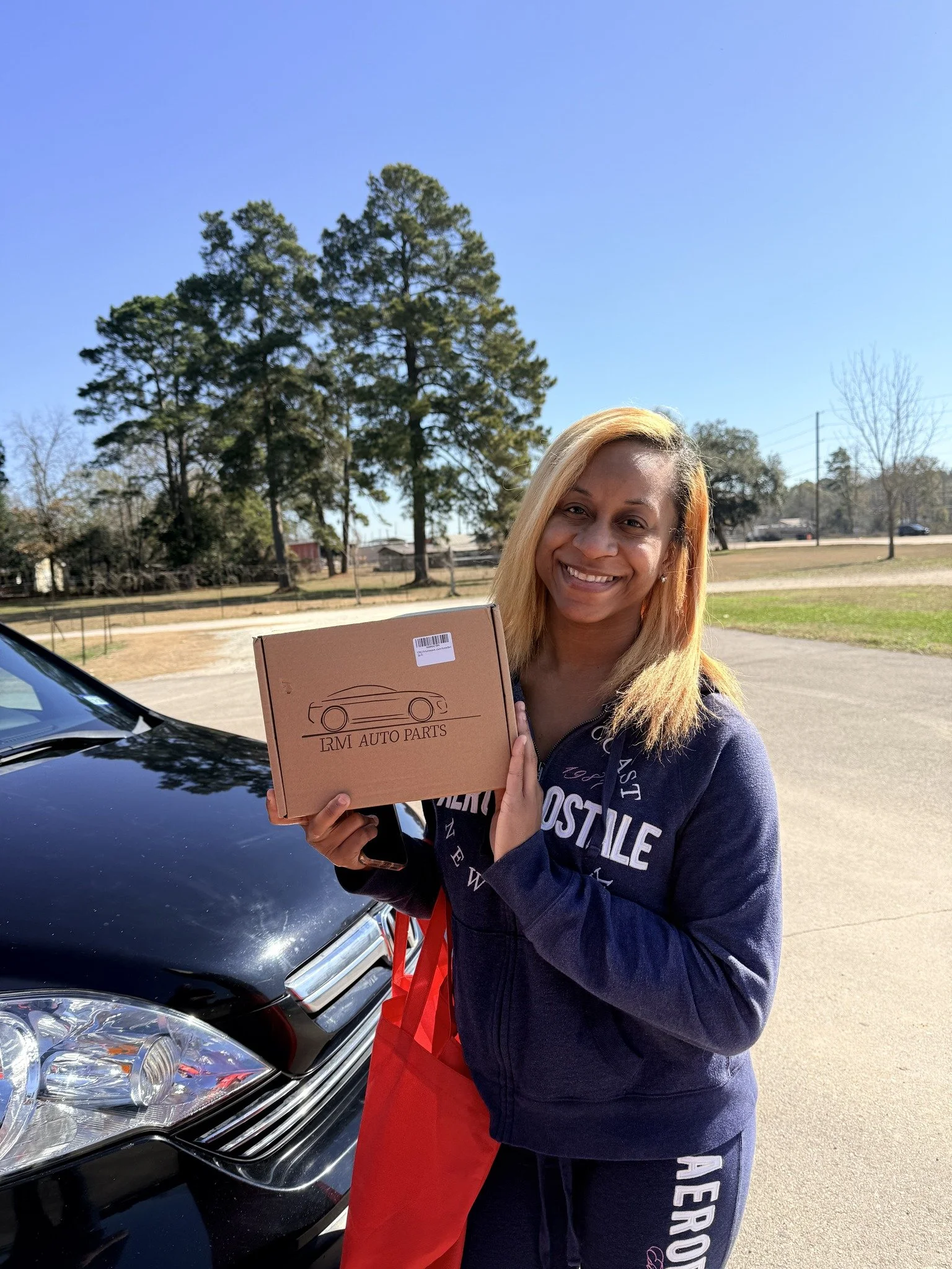 A woman with blonde hair smiling and holding a brown box labeled 'IRM Auto Parts' outdoors, standing next to a black car on a sunny day with dry grass and trees in the background.